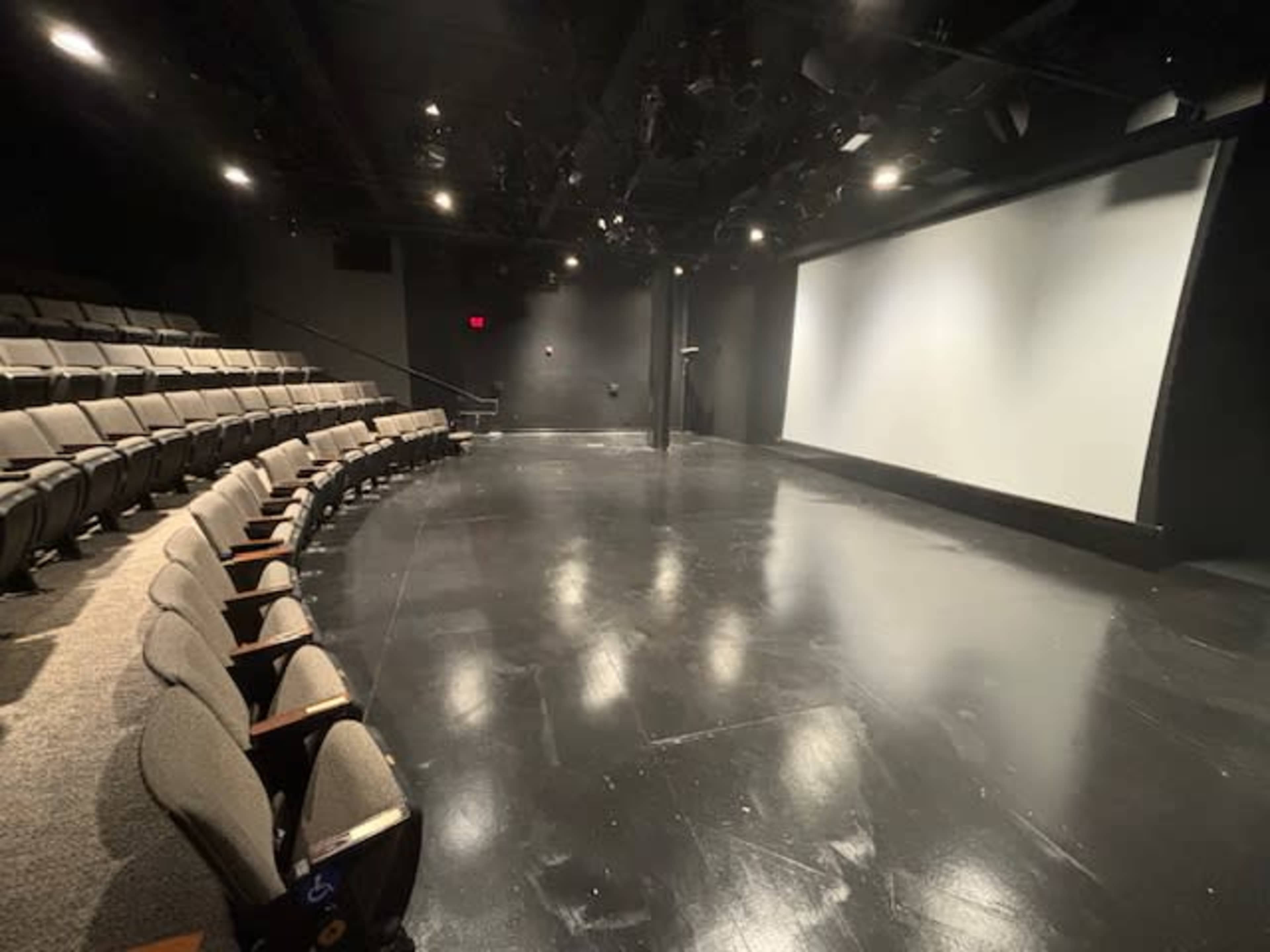 An empty black box theater with tiered seating and a large blank screen at the front.