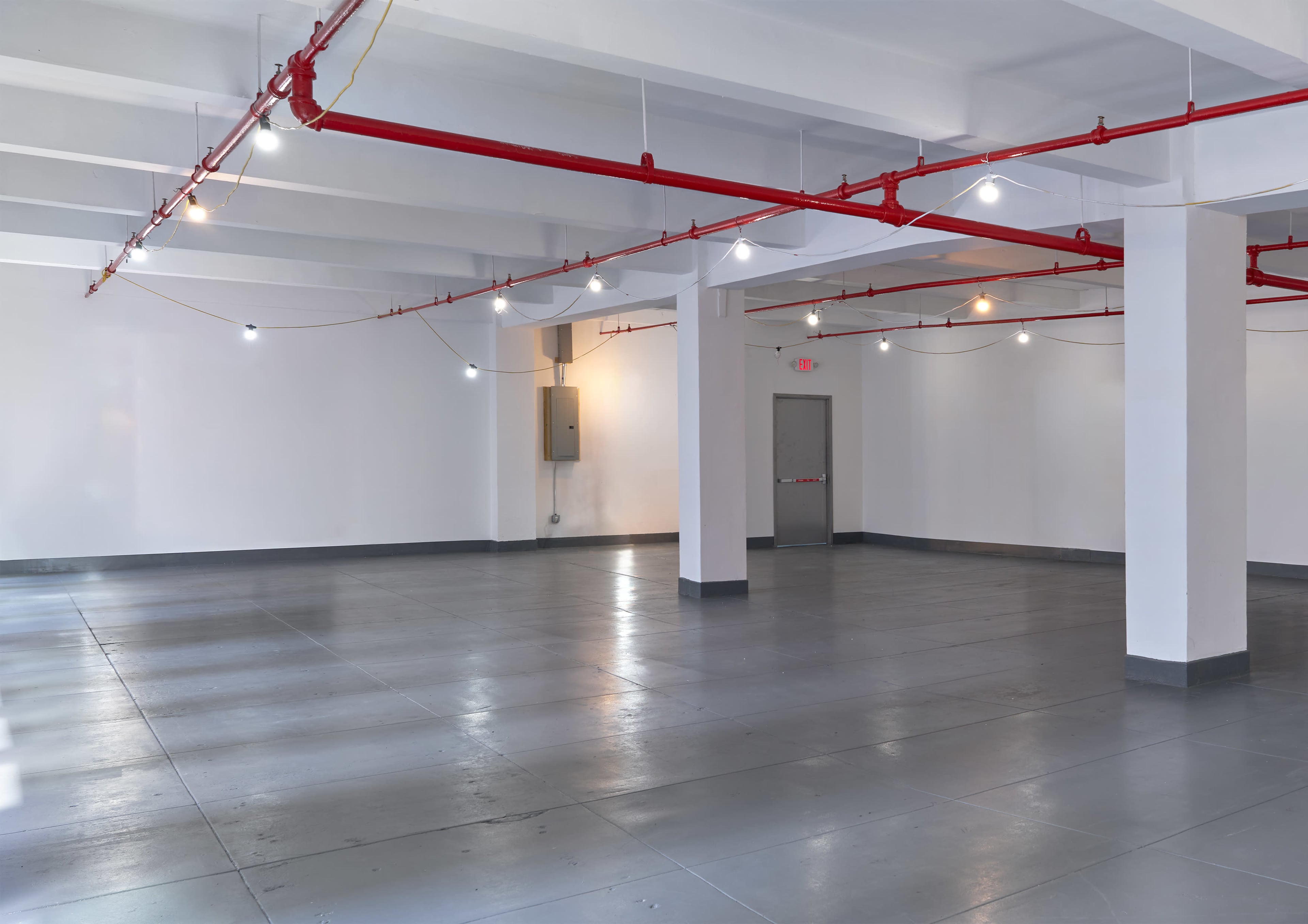 The image shows a spacious, empty room with polished gray flooring, white walls, and exposed red piping along the ceiling.