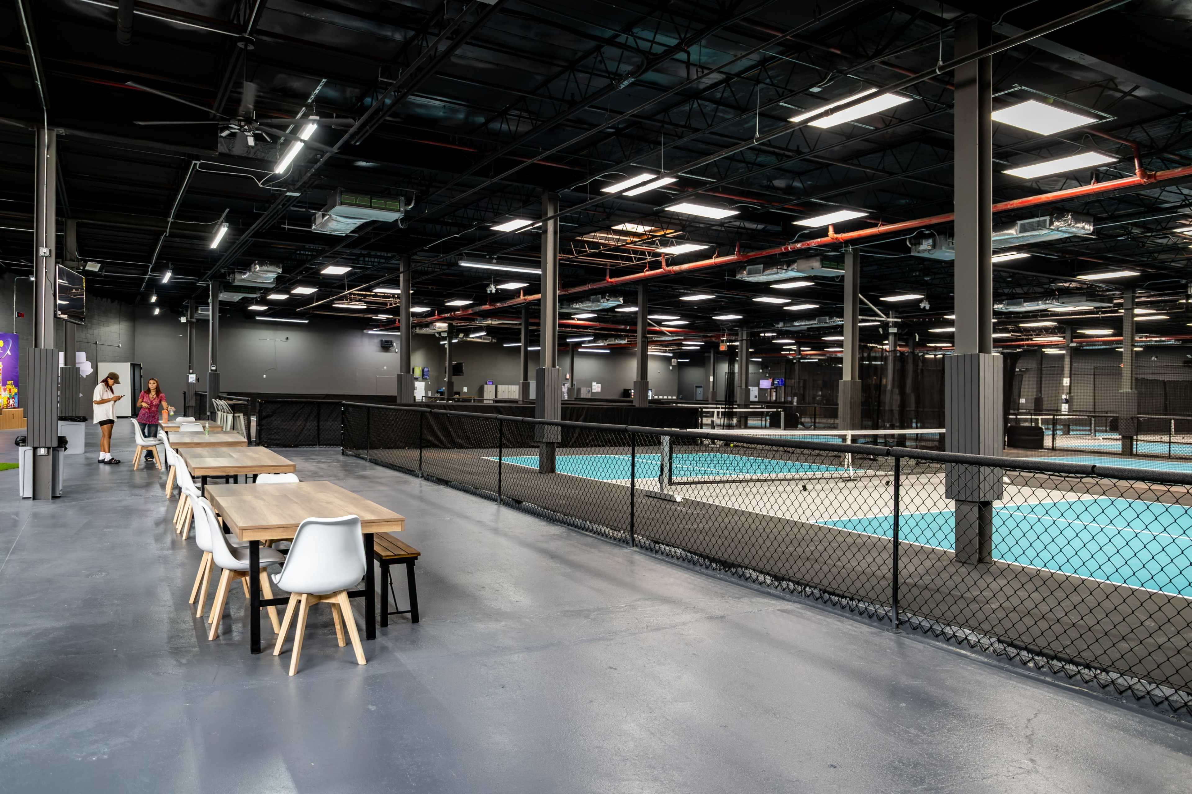The image shows a spacious indoor sports facility with several courts, surrounded by a dining area featuring tables and chairs.