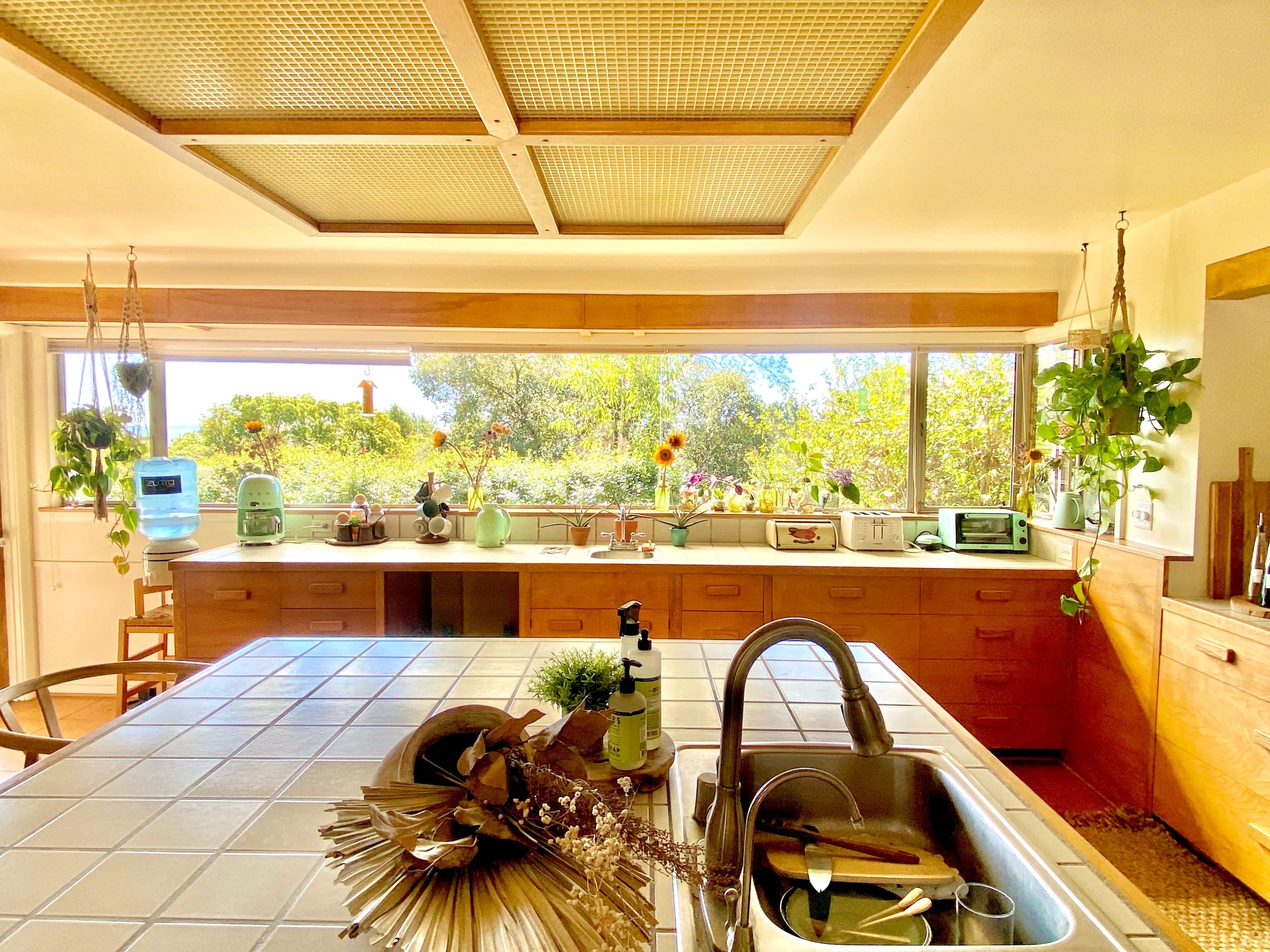 A bright kitchen features a tiled countertop, wooden cabinetry, and large windows showcasing greenery outside.