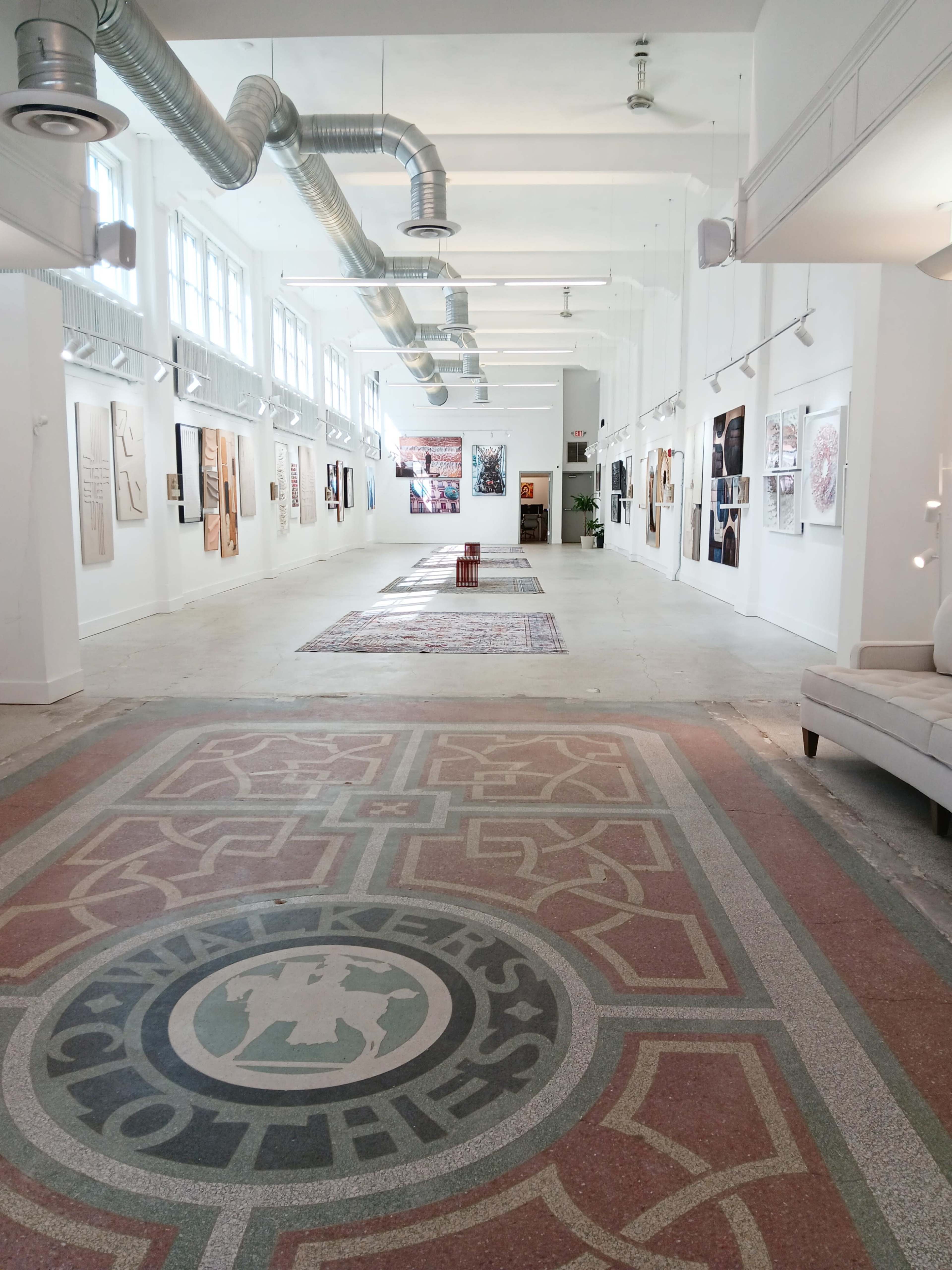 The image shows an art gallery with a spacious interior adorned with various artworks displayed along the walls and a patterned floor.