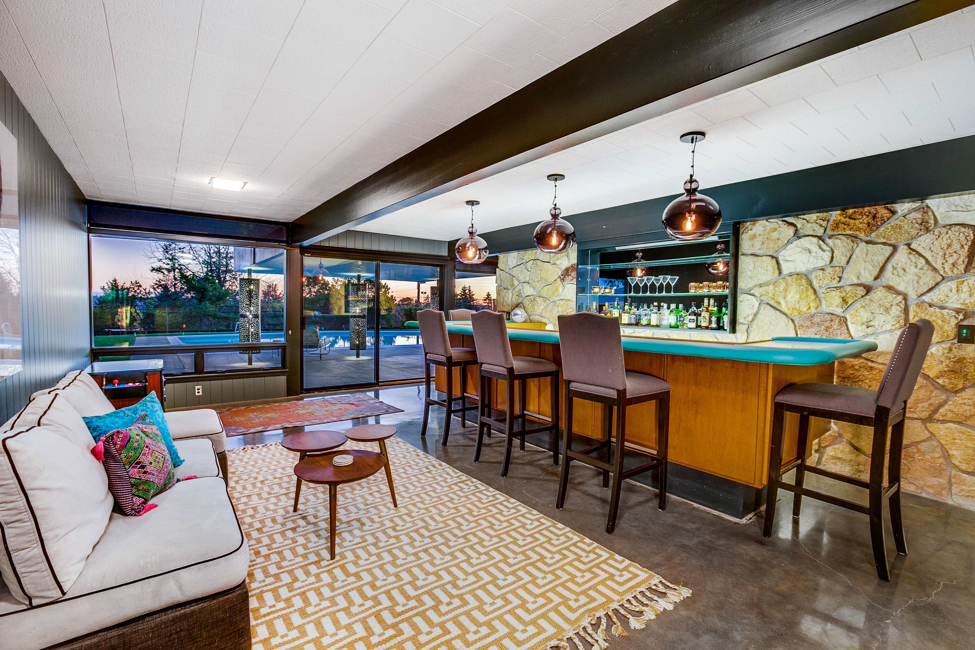 A modern home basement features a stone bar area with high stools, a couch, and large windows overlooking a pool and garden.