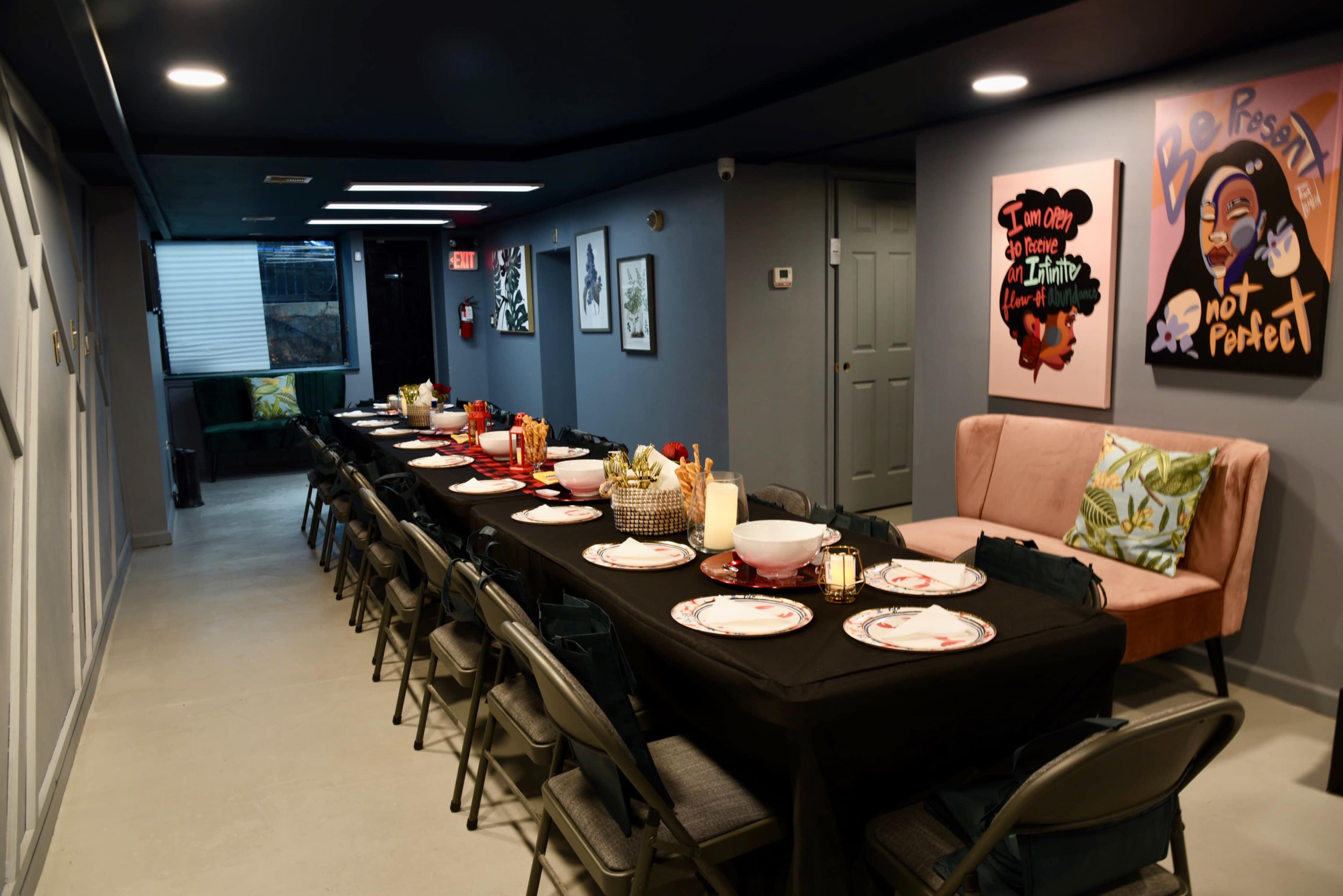 A long table is set for a meal, decorated with plates and glasses, and surrounded by folding chairs in a dimly lit room with artwork on the walls.