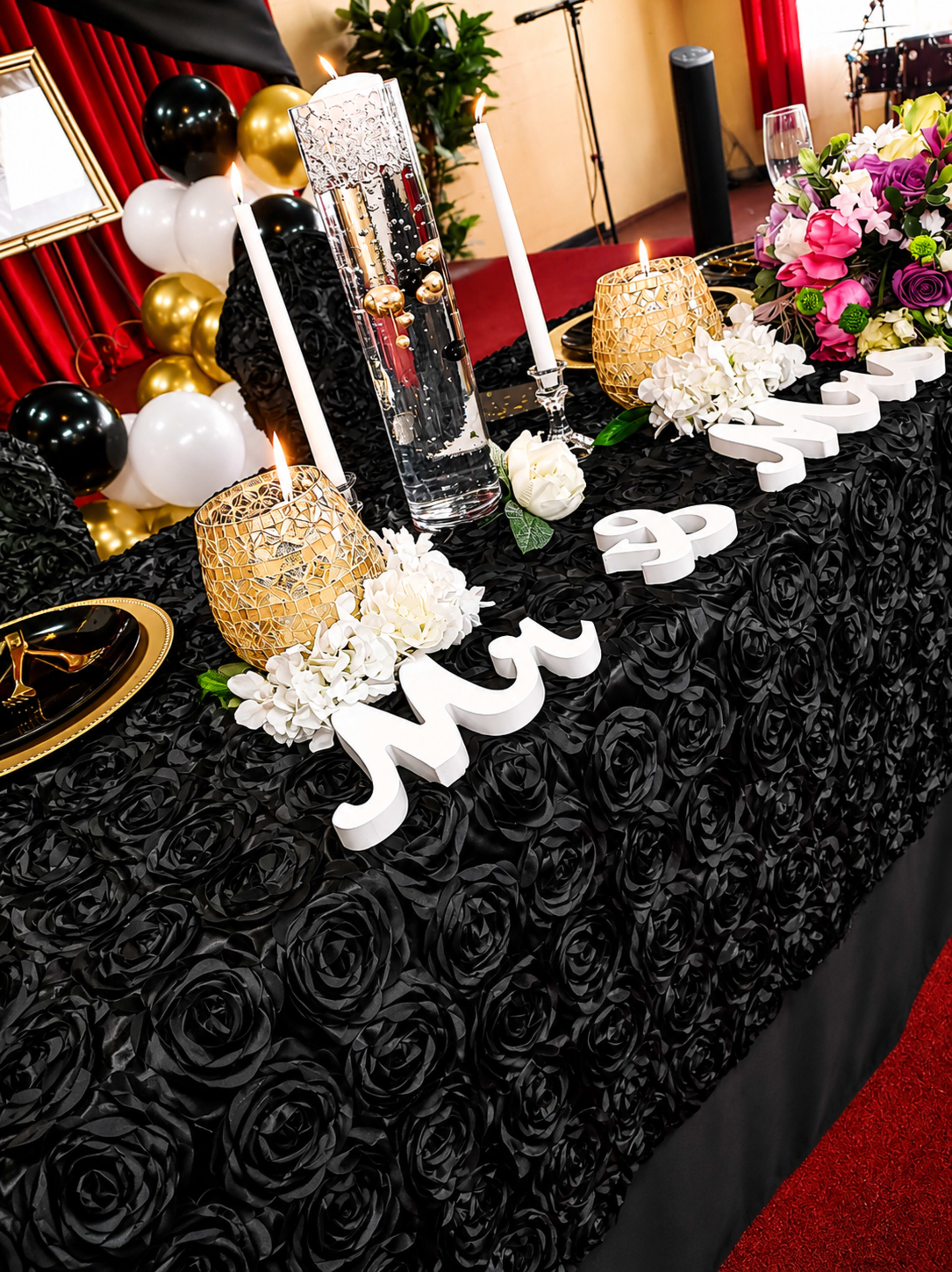 A decorated table is set with white floral arrangements, candles, and letters spelling "Mr & Mrs," all against a black rose-patterned tablecloth.