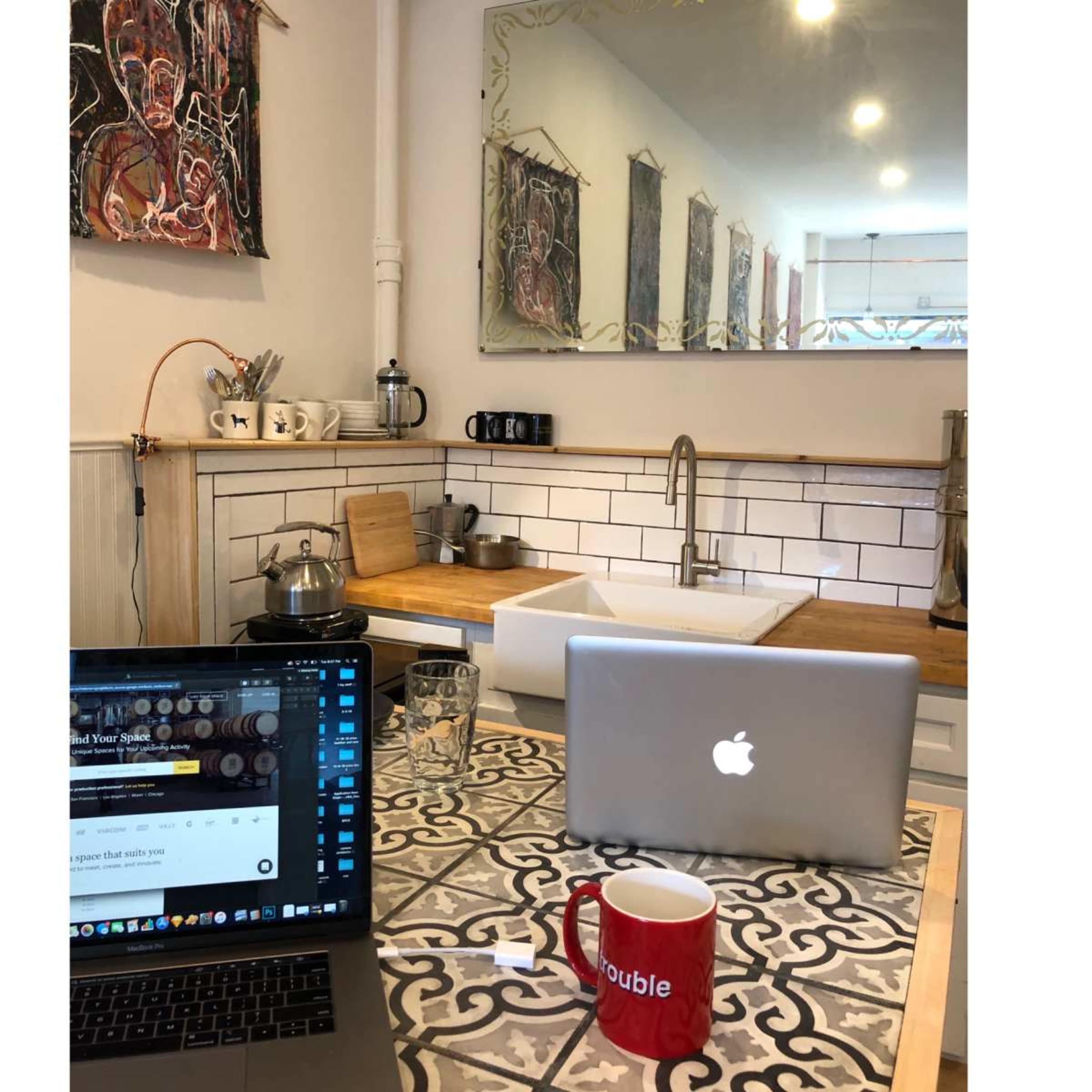 A kitchen space with a wooden countertop, sink, and two laptops on a patterned table, surrounded by kitchen utensils and artwork on the walls.