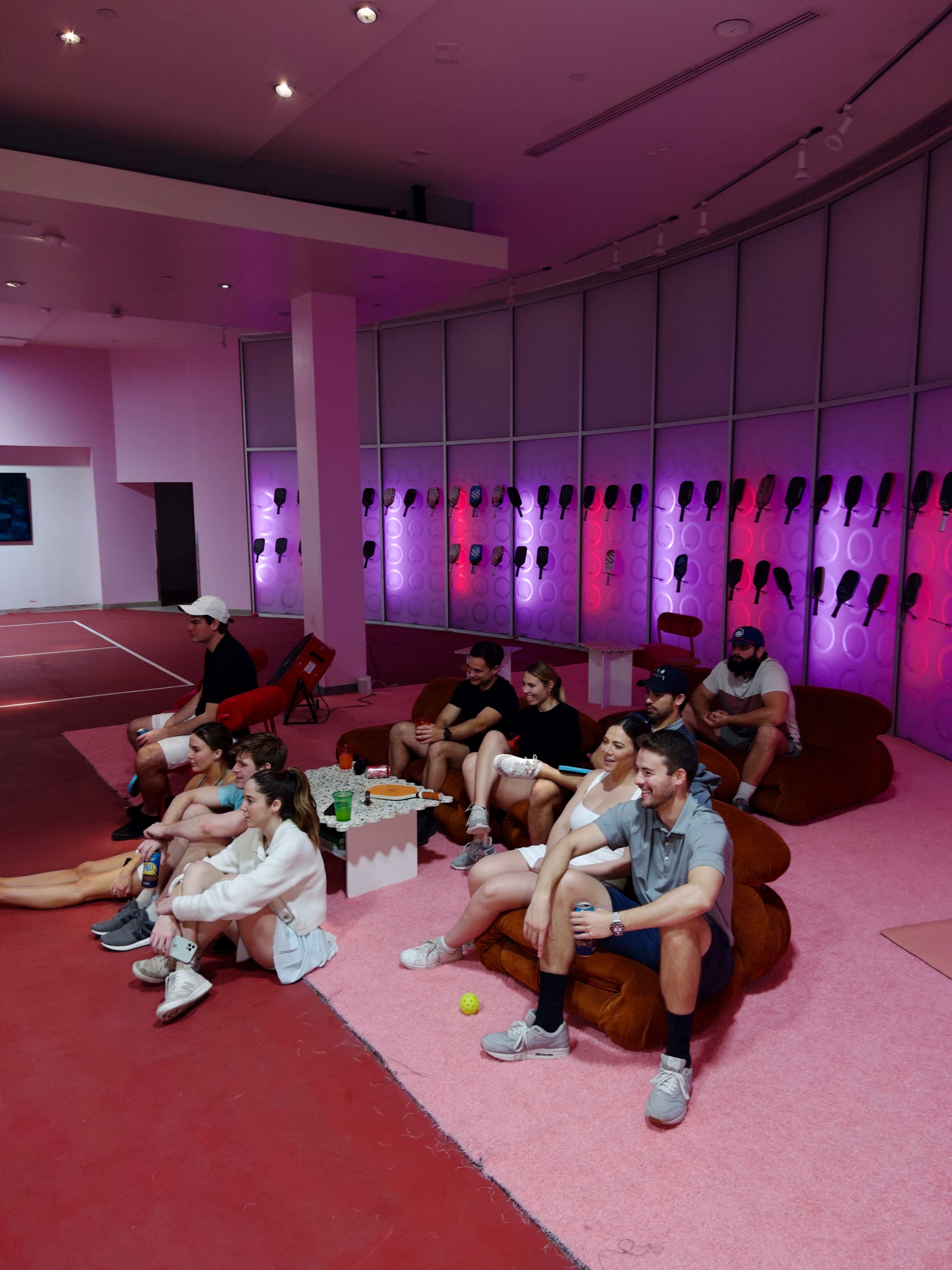 A group of people sits on large cushions in a brightly lit room with pink walls and colored lighting, while others play tennis in the background.