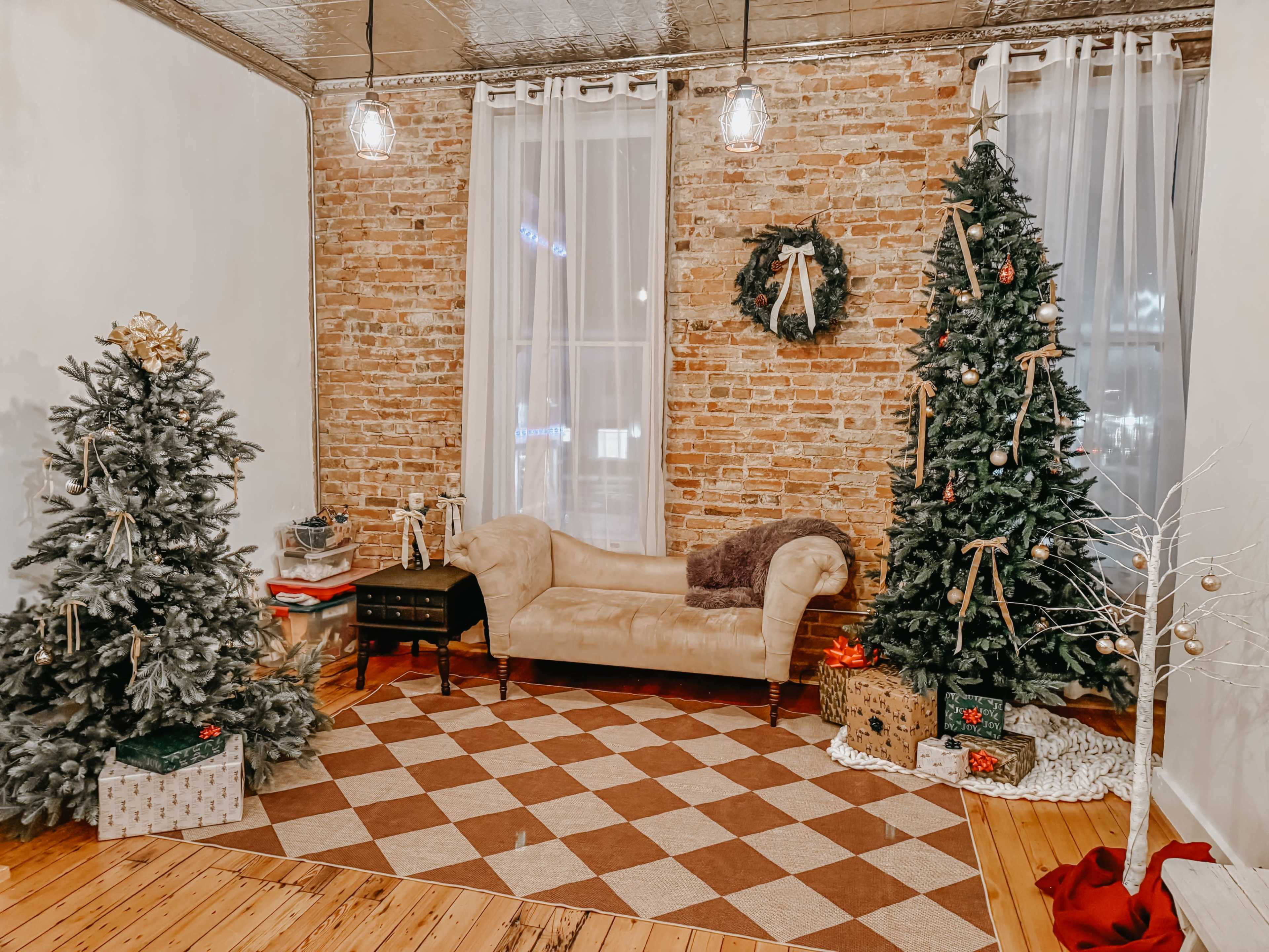 The image shows a cozy room decorated for Christmas, featuring two Christmas trees, a couch, a checkered rug, and festive decorations against a brick wall.