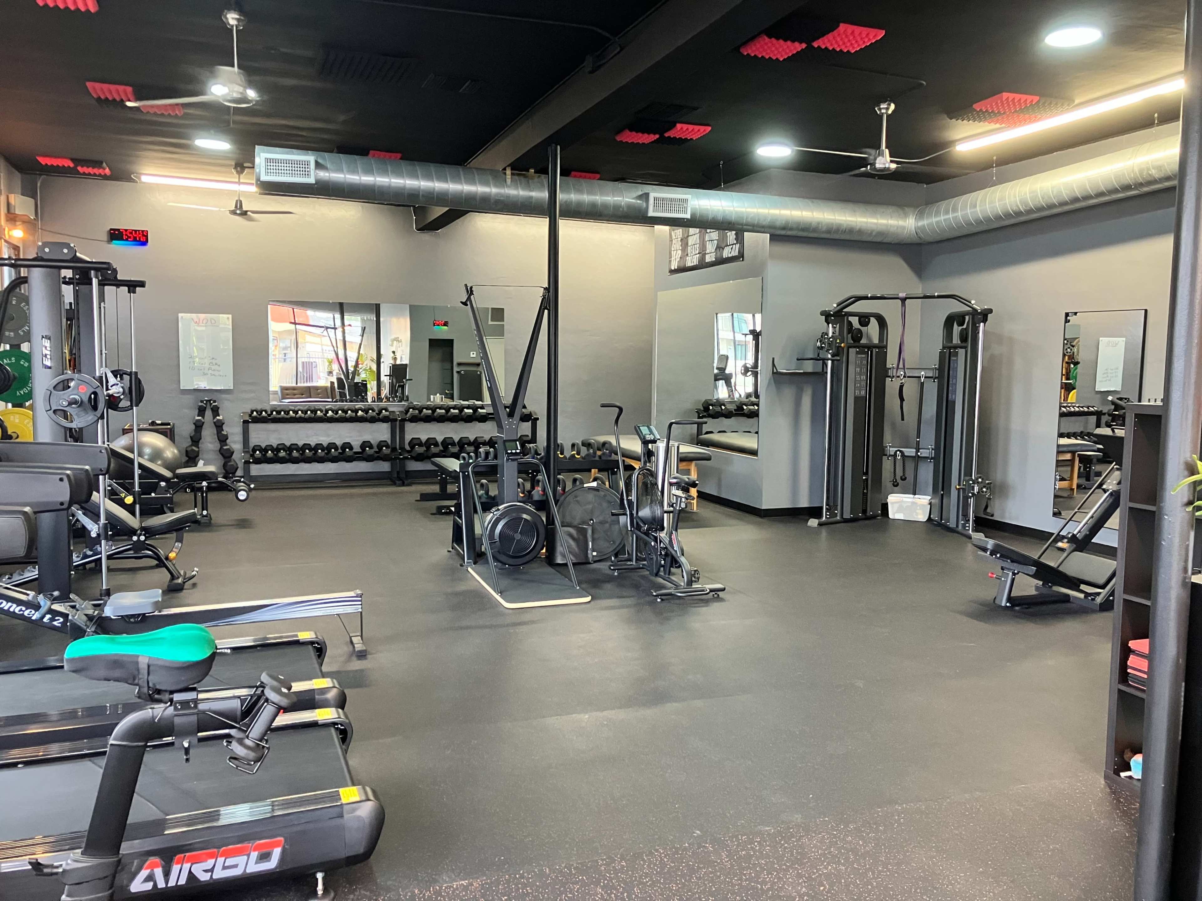 The image shows an indoor gym with various exercise equipment, including weight machines, free weights, and cardio machines, arranged neatly in a spacious area.
