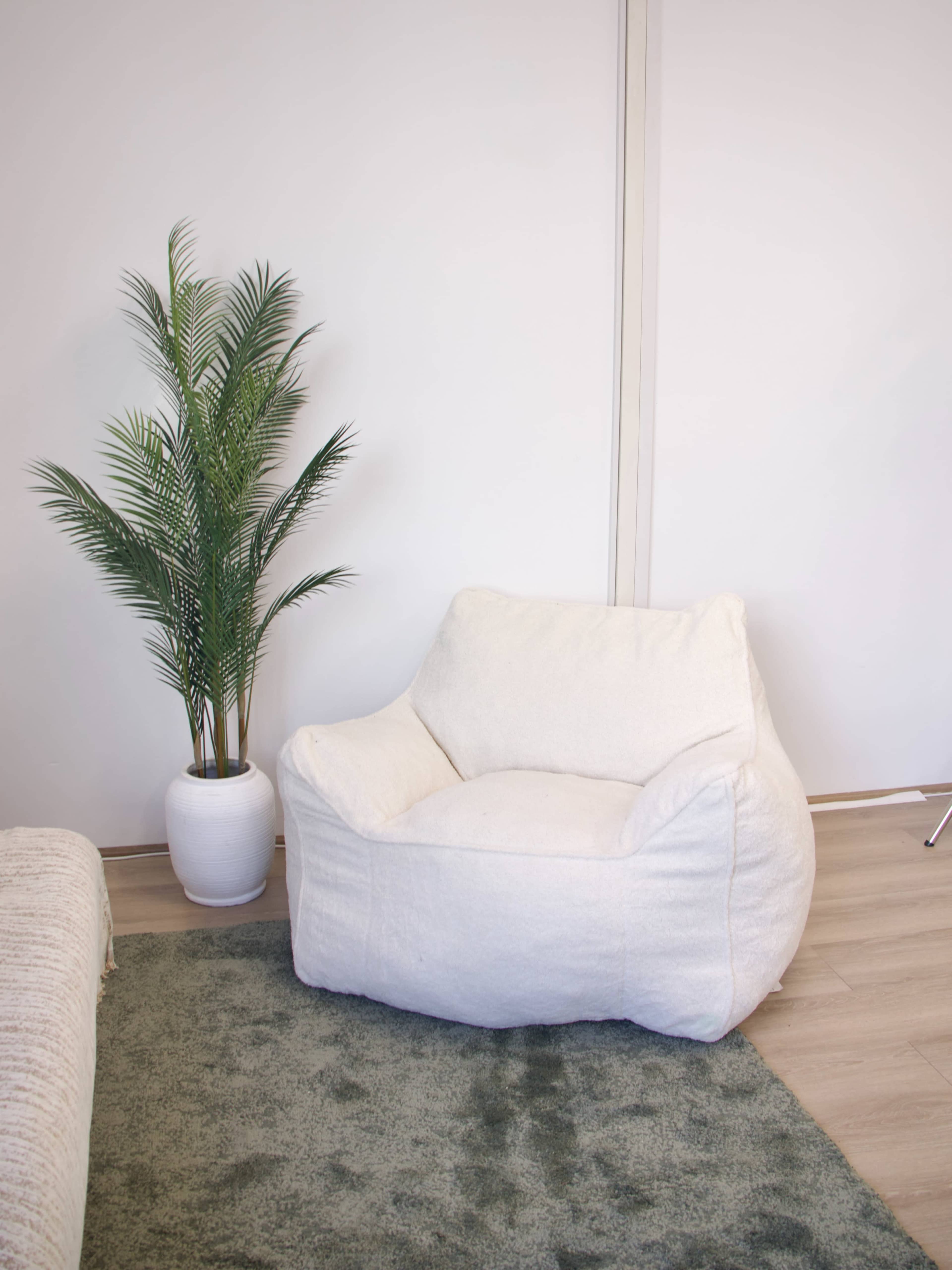 The image shows a white bean bag chair next to a potted palm plant on a green rug in a minimalist room.