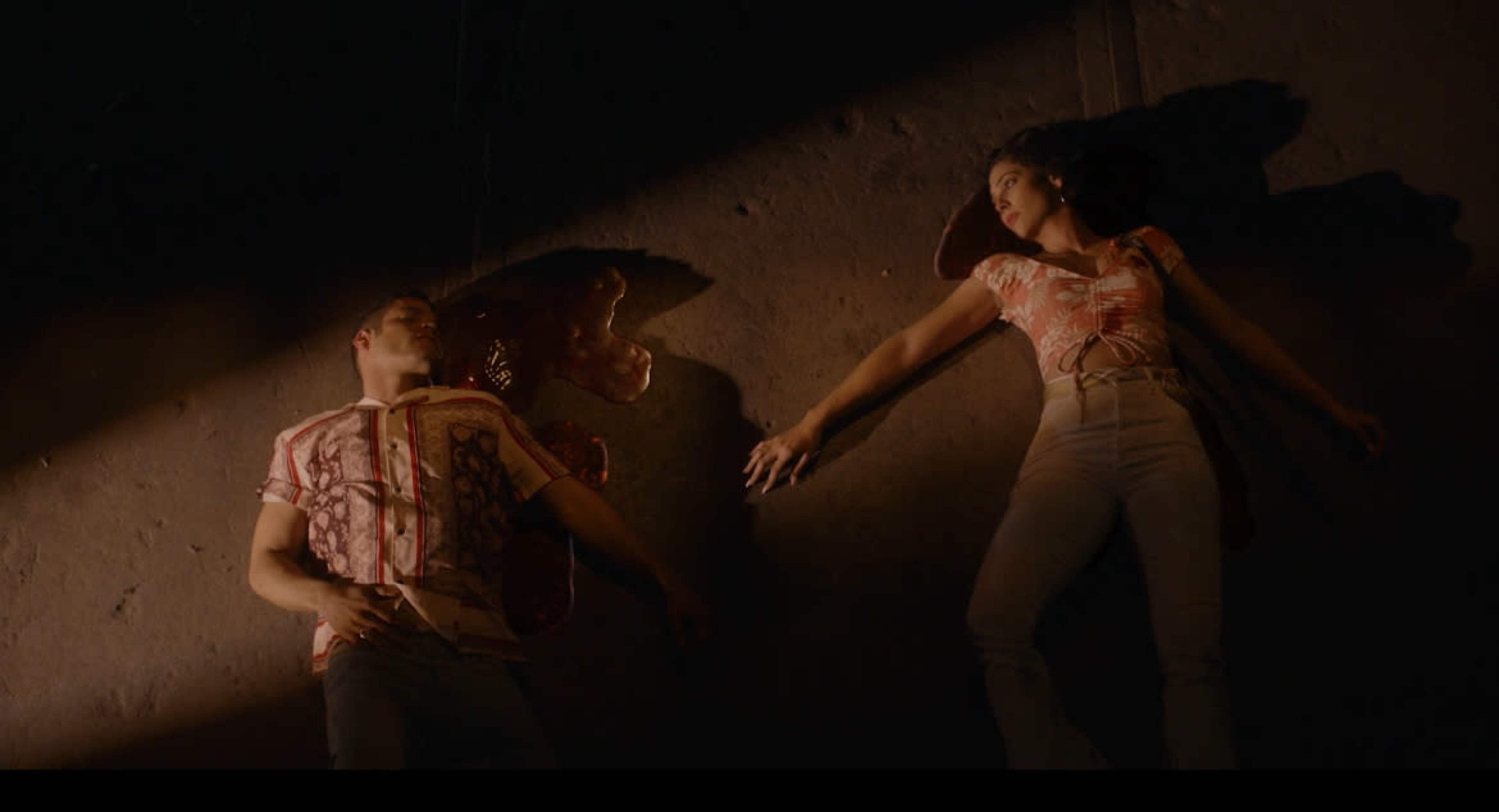 A man and a woman lie on the ground in a dimly lit space, with a pool of blood near the man.
