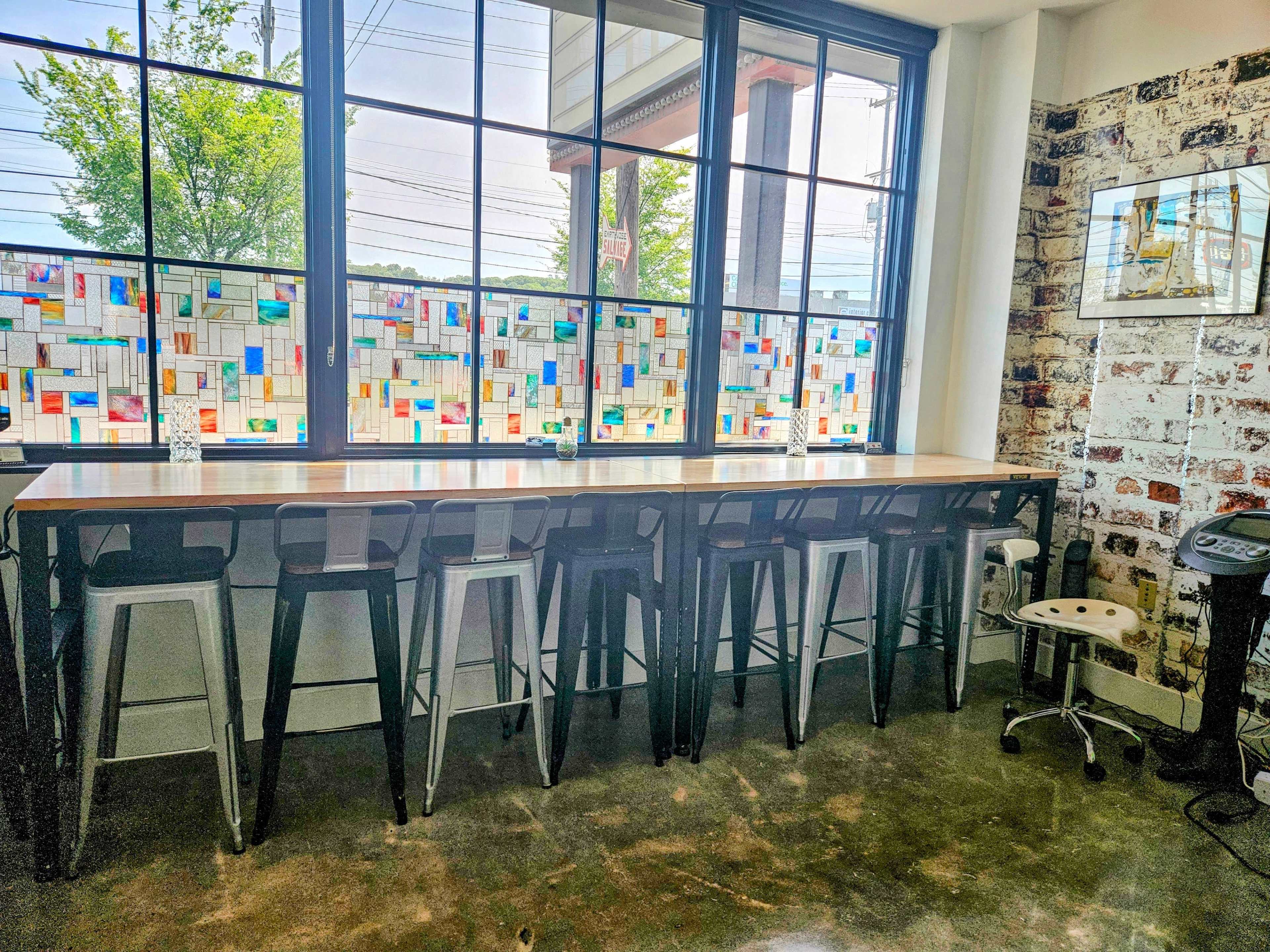 A modern cafe interior with a long wooden counter and metal stools, backed by colorful stained glass windows and a brick wall.