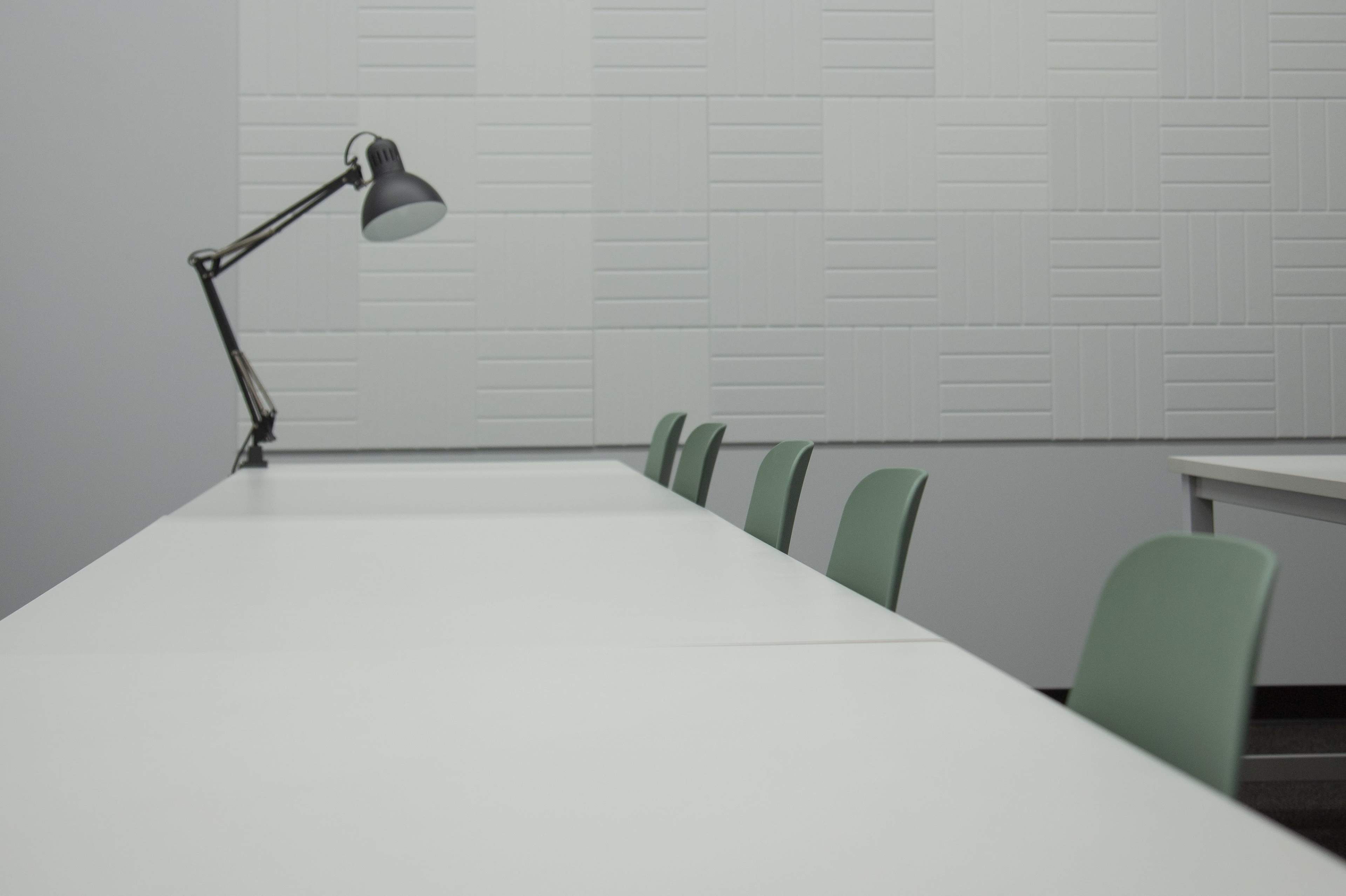 A row of green chairs sits at a long, white table in a minimalist room with a desk lamp and textured wall.