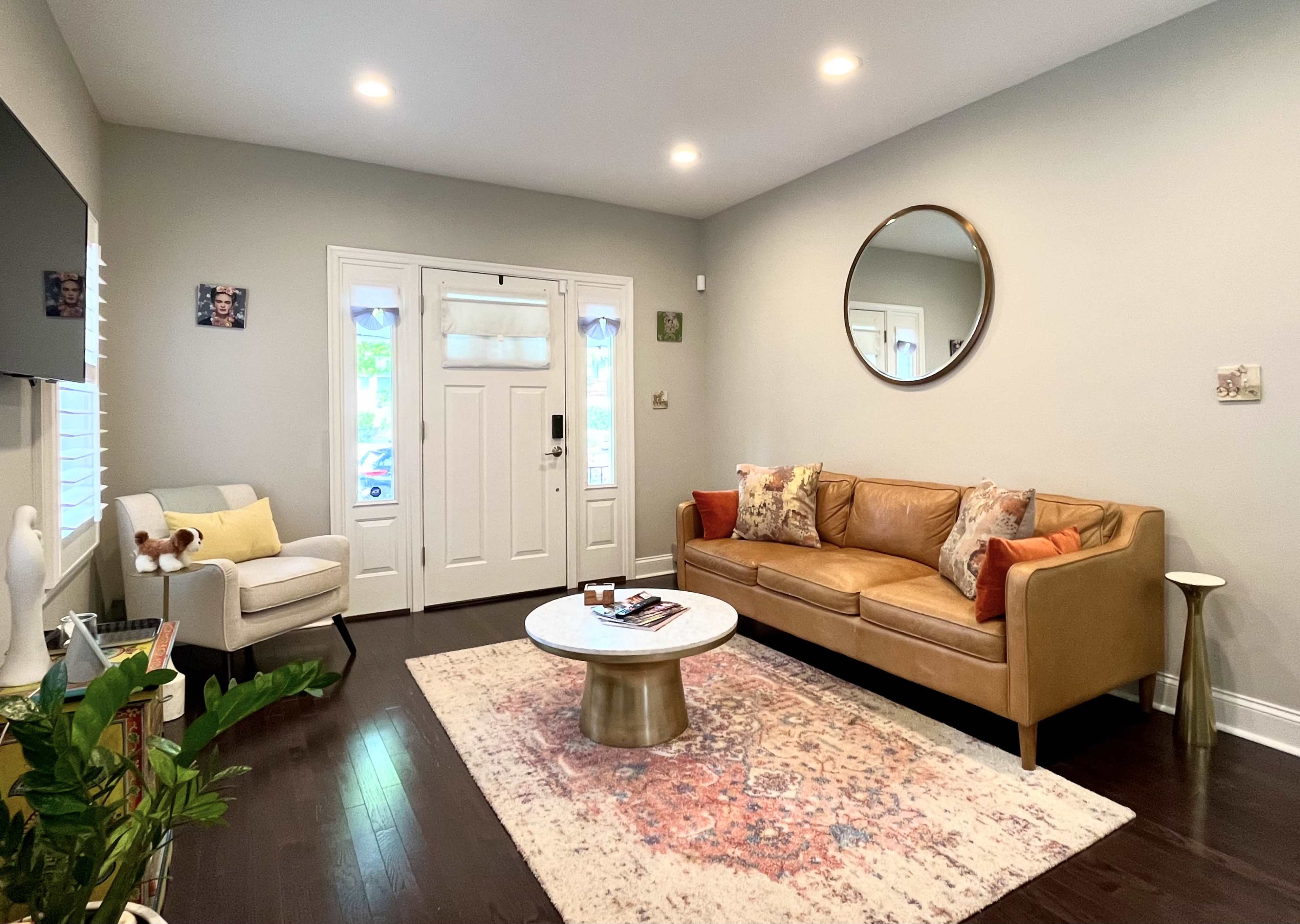 A cozy living room features a brown leather couch, a circular mirror, a round coffee table, and a light-colored rug, with a front door visible in the background.
