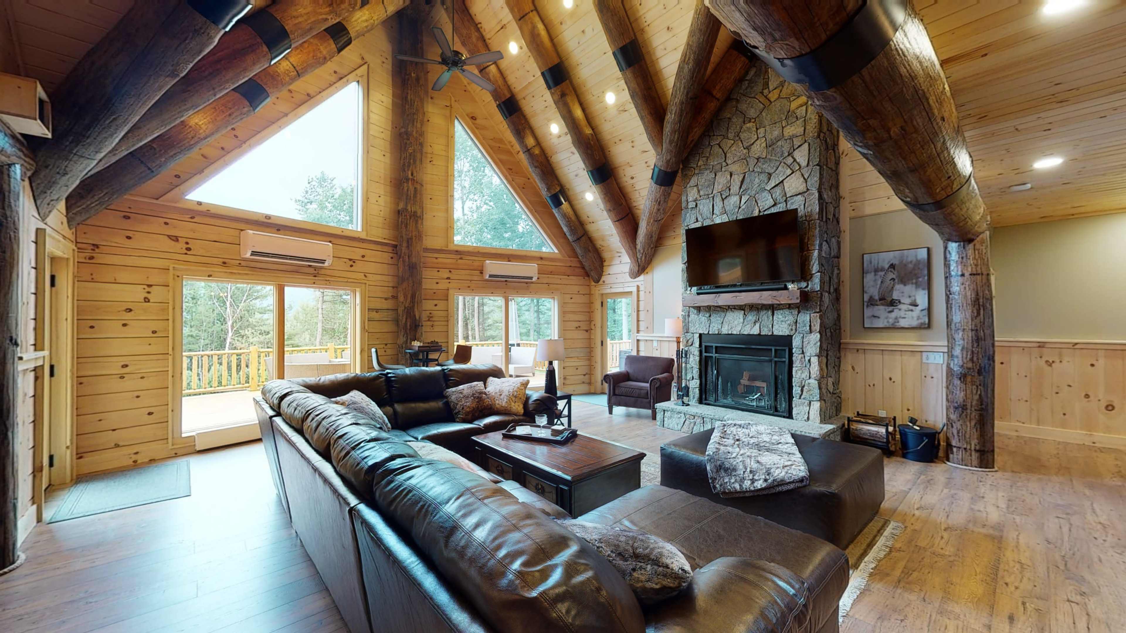 A spacious wooden living room with a stone fireplace, large windows, and vaulted ceilings supported by exposed beams.