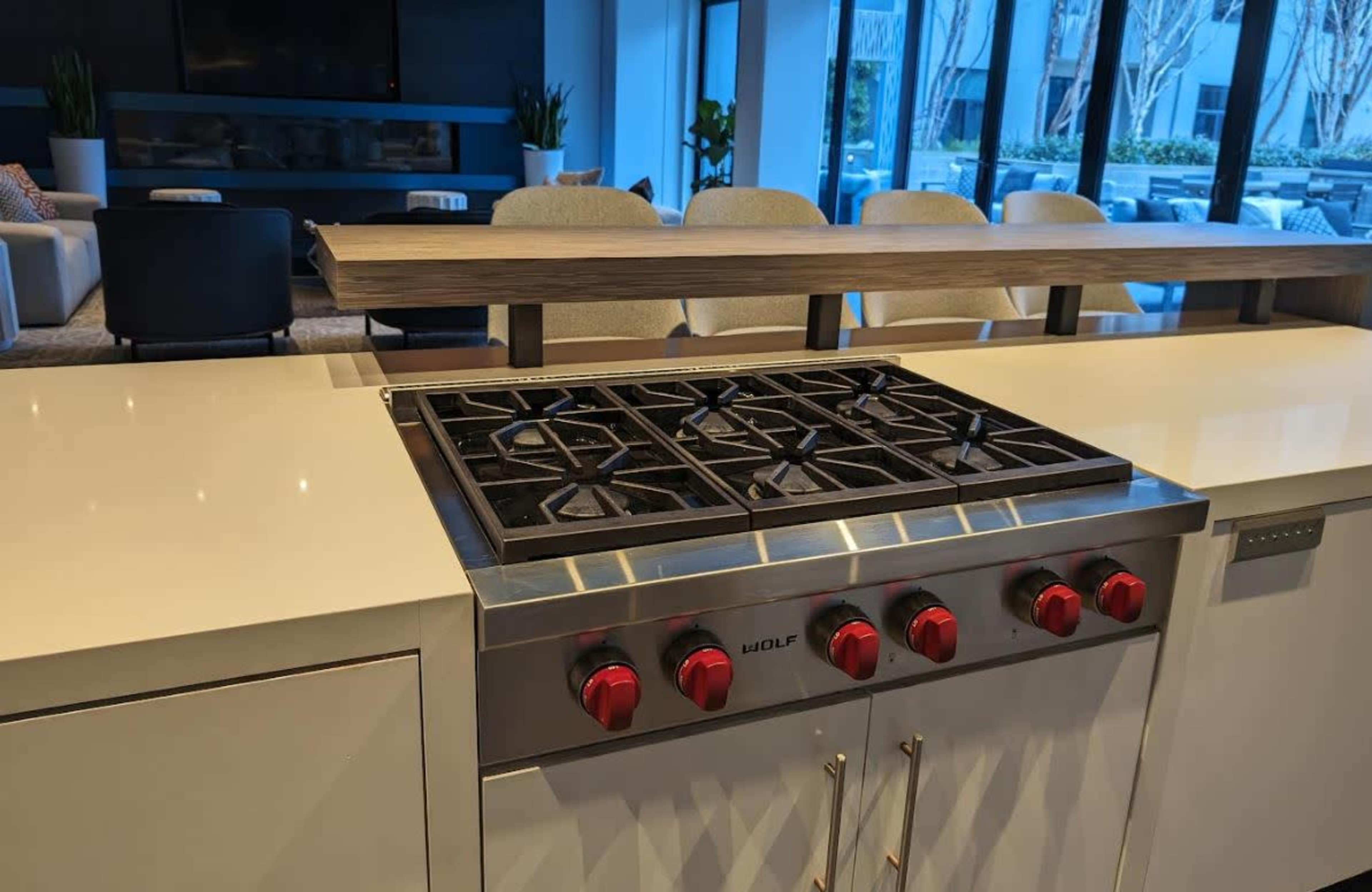 A modern kitchen with a stainless steel Wolf gas cooktop featuring five burners and red knobs, set against a backdrop of a brightly lit living area.