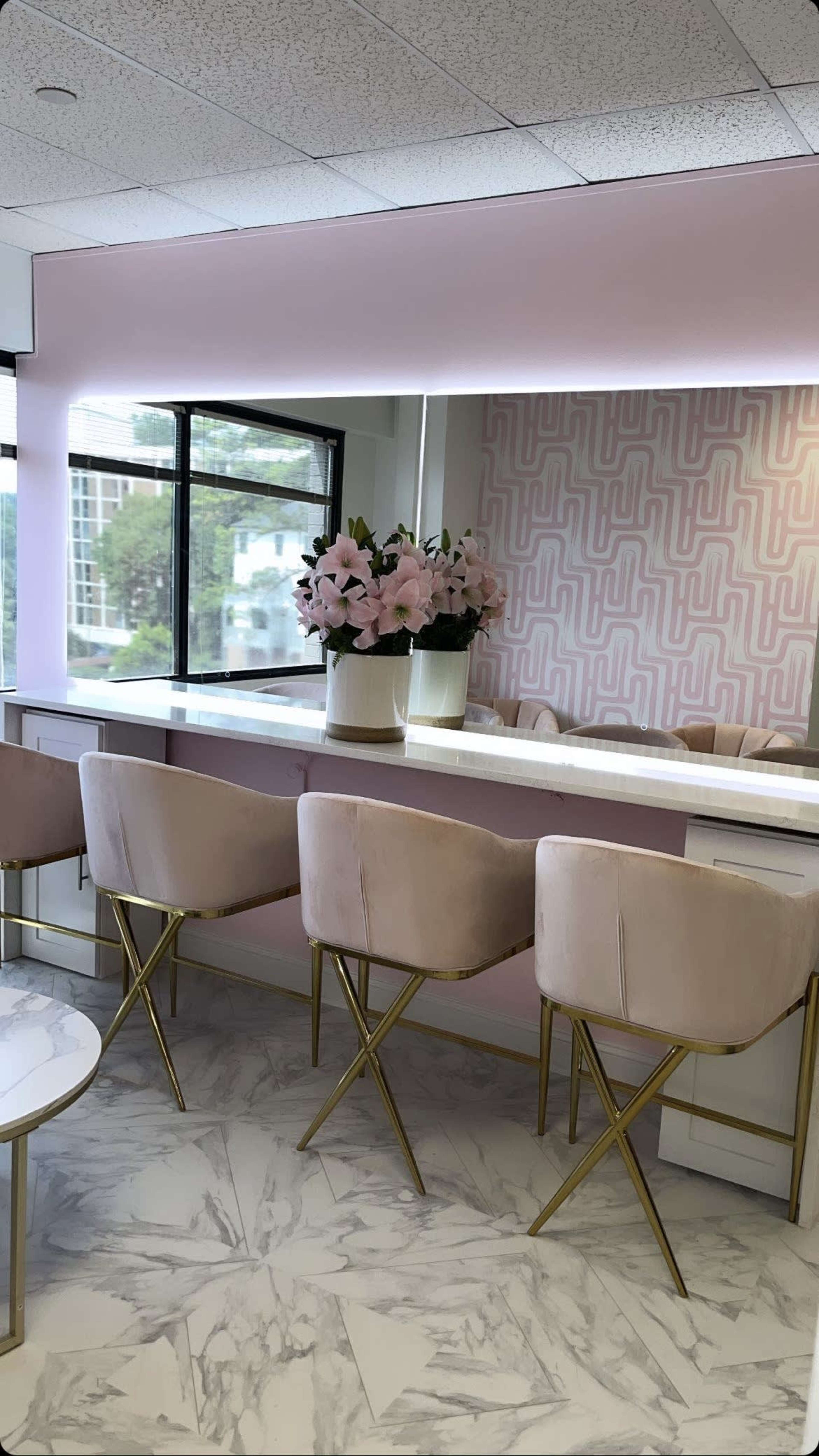 A modern interior with a light pink accent wall, marble flooring, and several upholstered chairs arranged around a counter with a floral centerpiece.