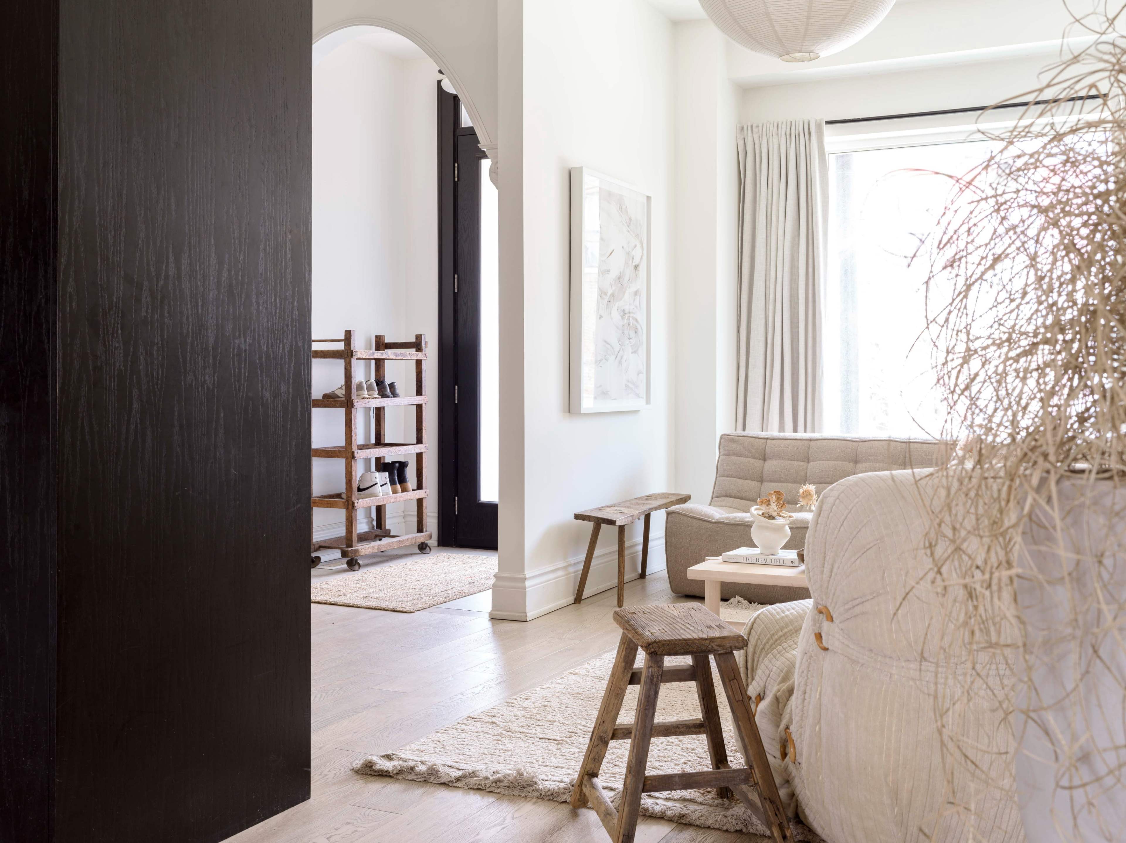 A modern living room with a neutral color palette, featuring a sofa, a small wooden table, and a glimpse of a hallway with a wooden shelving unit.