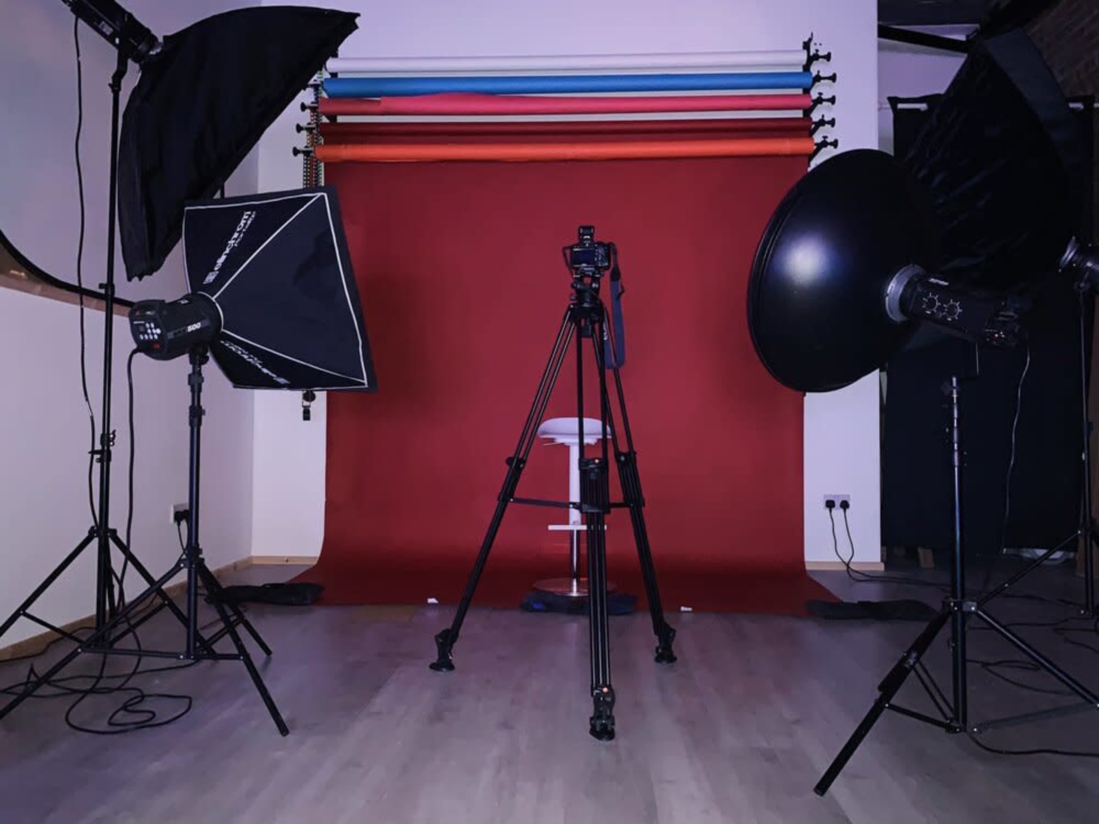 A photography studio set up with a red backdrop, a camera on a tripod, and several lighting equipment positioned around a white stool.