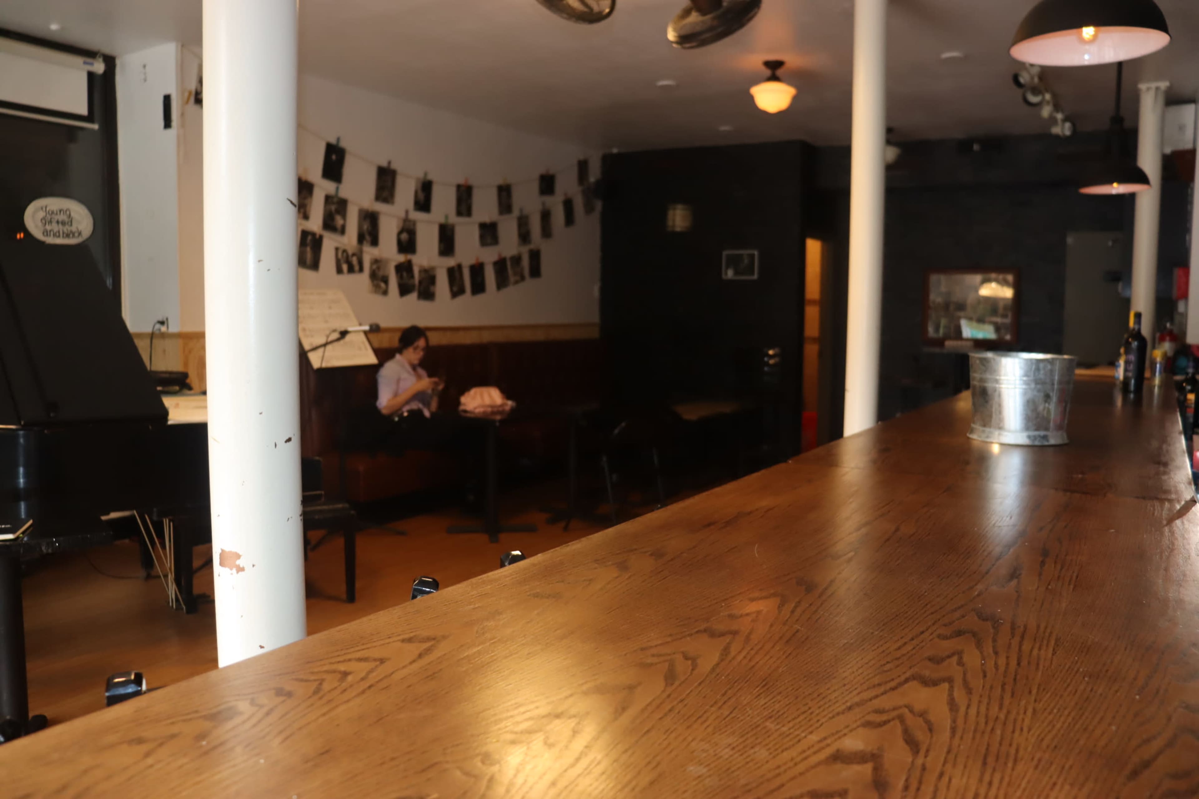 A bar with a polished wooden counter and scattered seating areas, featuring a woman sitting on her phone in the background.