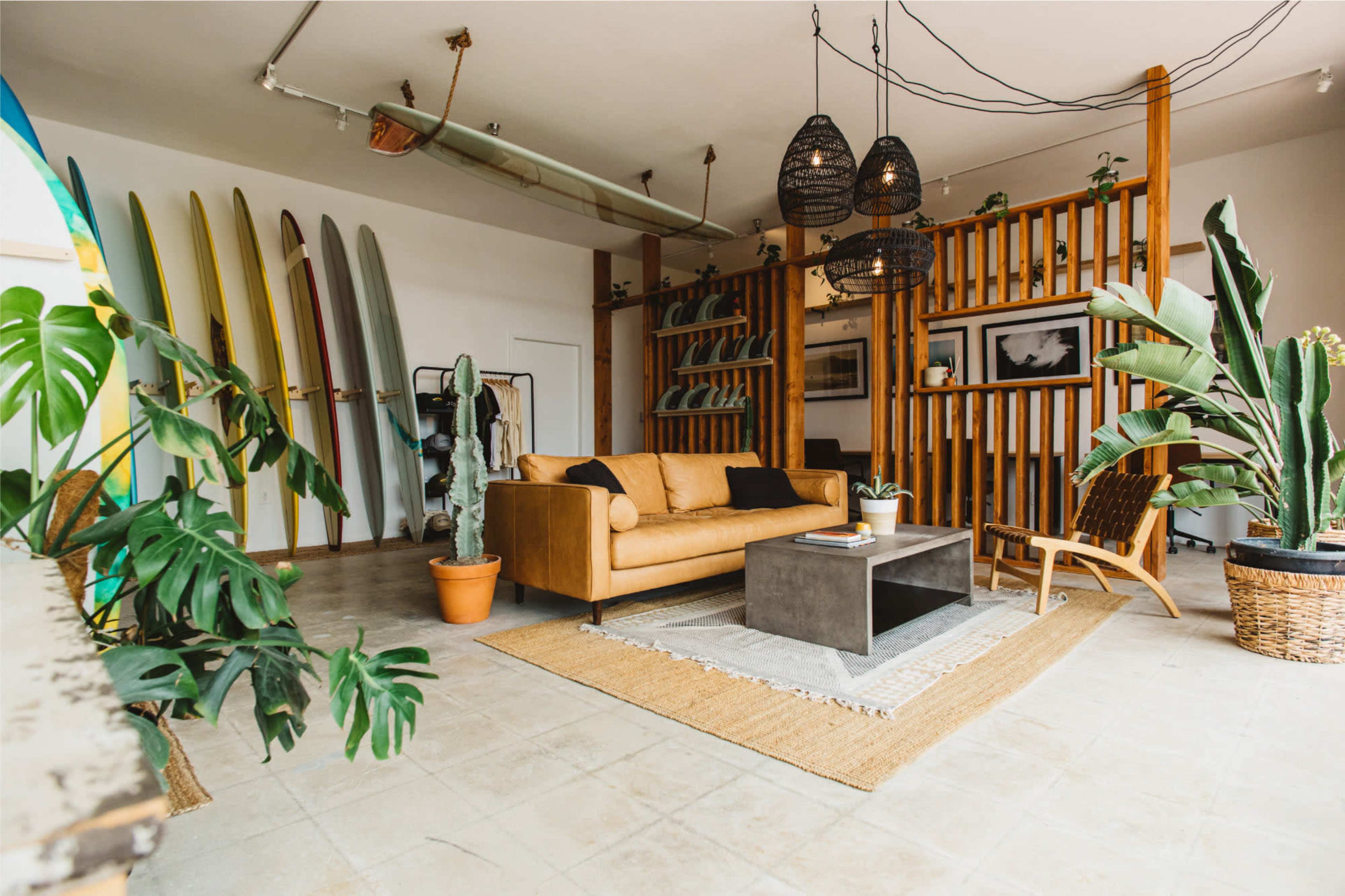 The image shows a modern living room featuring a tan sofa, a concrete coffee table, and decorative surfboards mounted on the wall, surrounded by plants and stylish lighting fixtures.