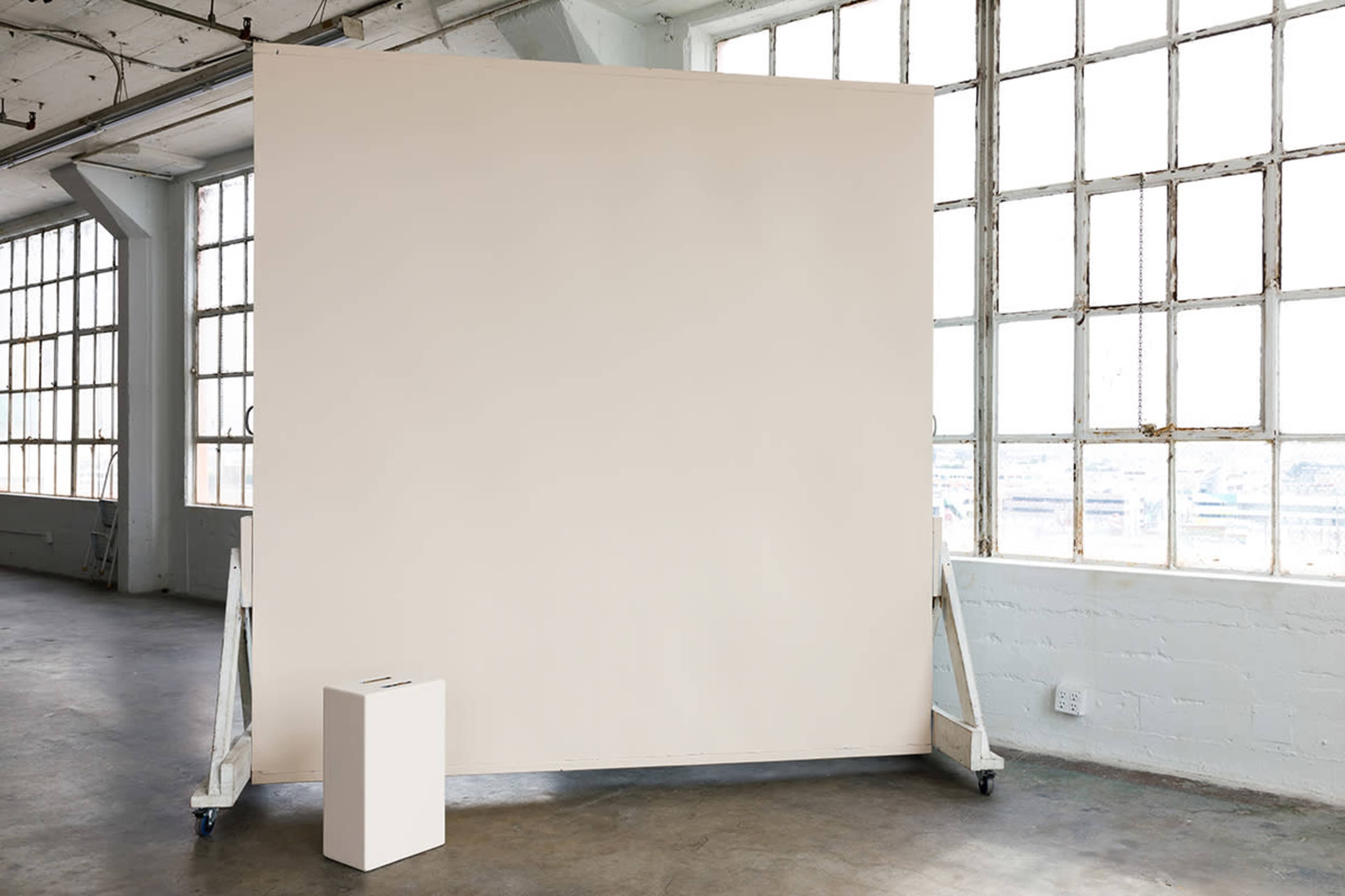 The image shows a large blank wall supported by a frame, near a white object on the floor in an industrial space with large windows.