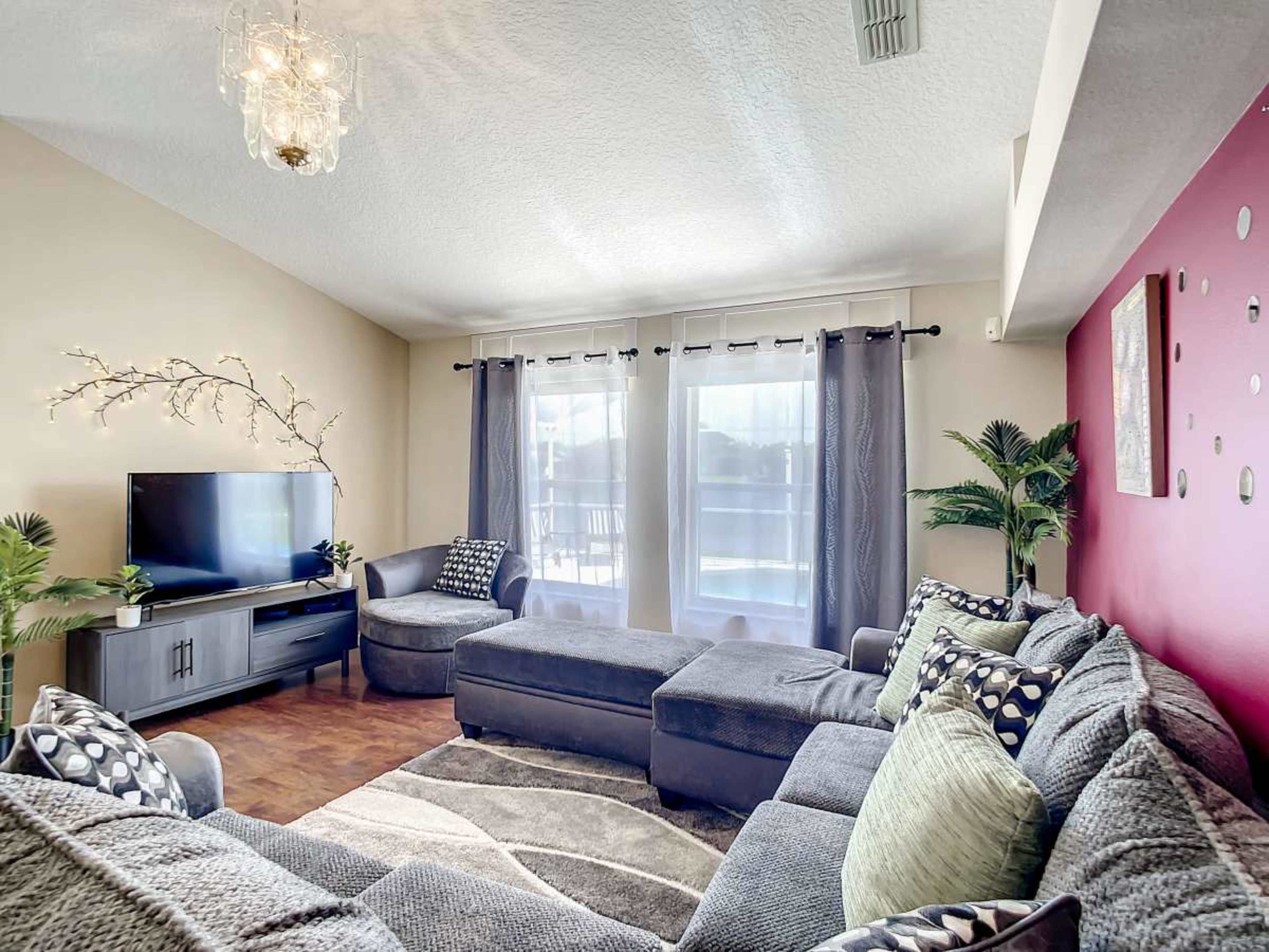 A modern living room features a sectional sofa, a television, and large windows with curtains.