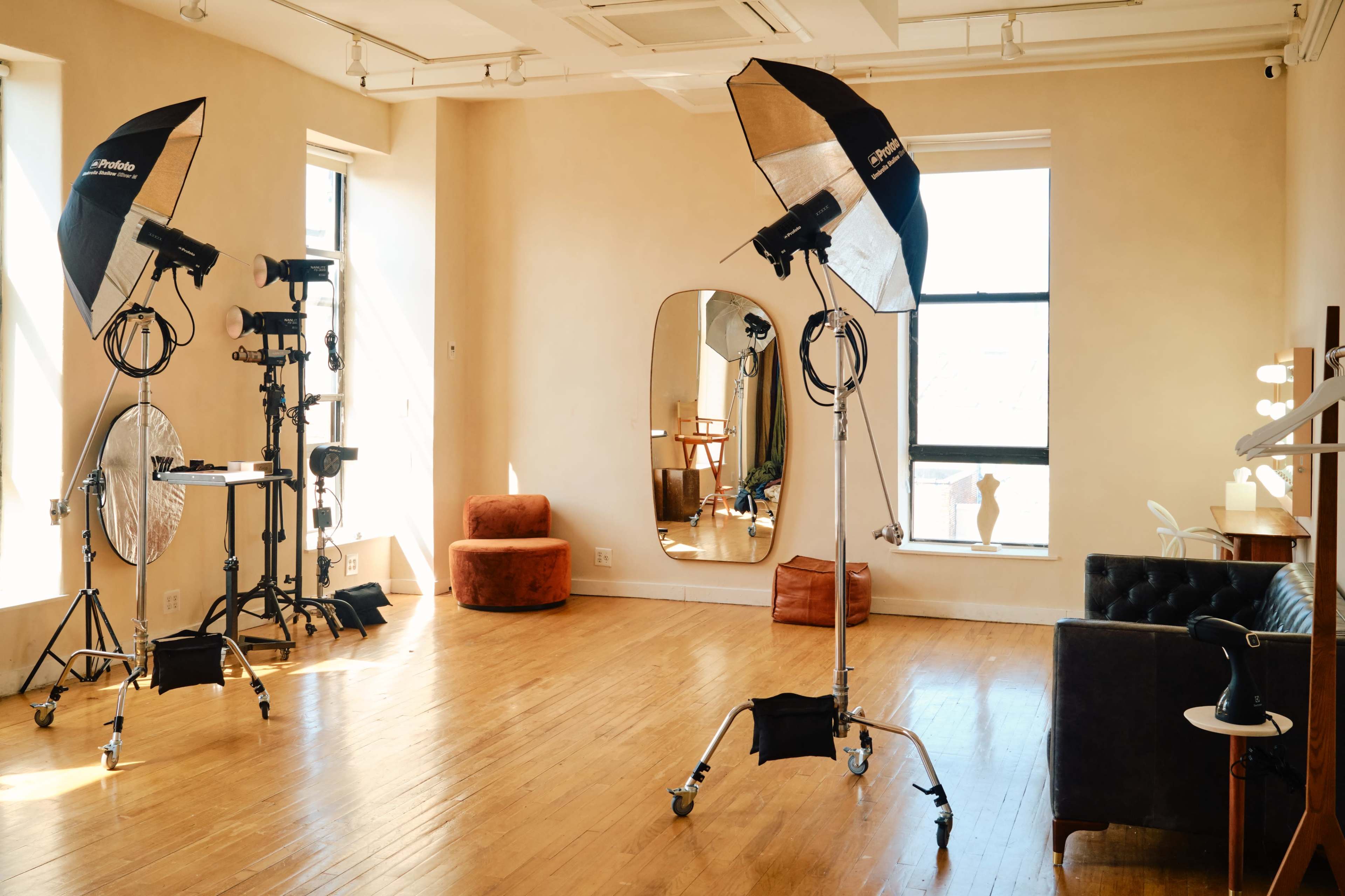 The image shows a well-lit photo studio with softbox lights, a large mirror, a brown couch, and a few props arranged in the space.