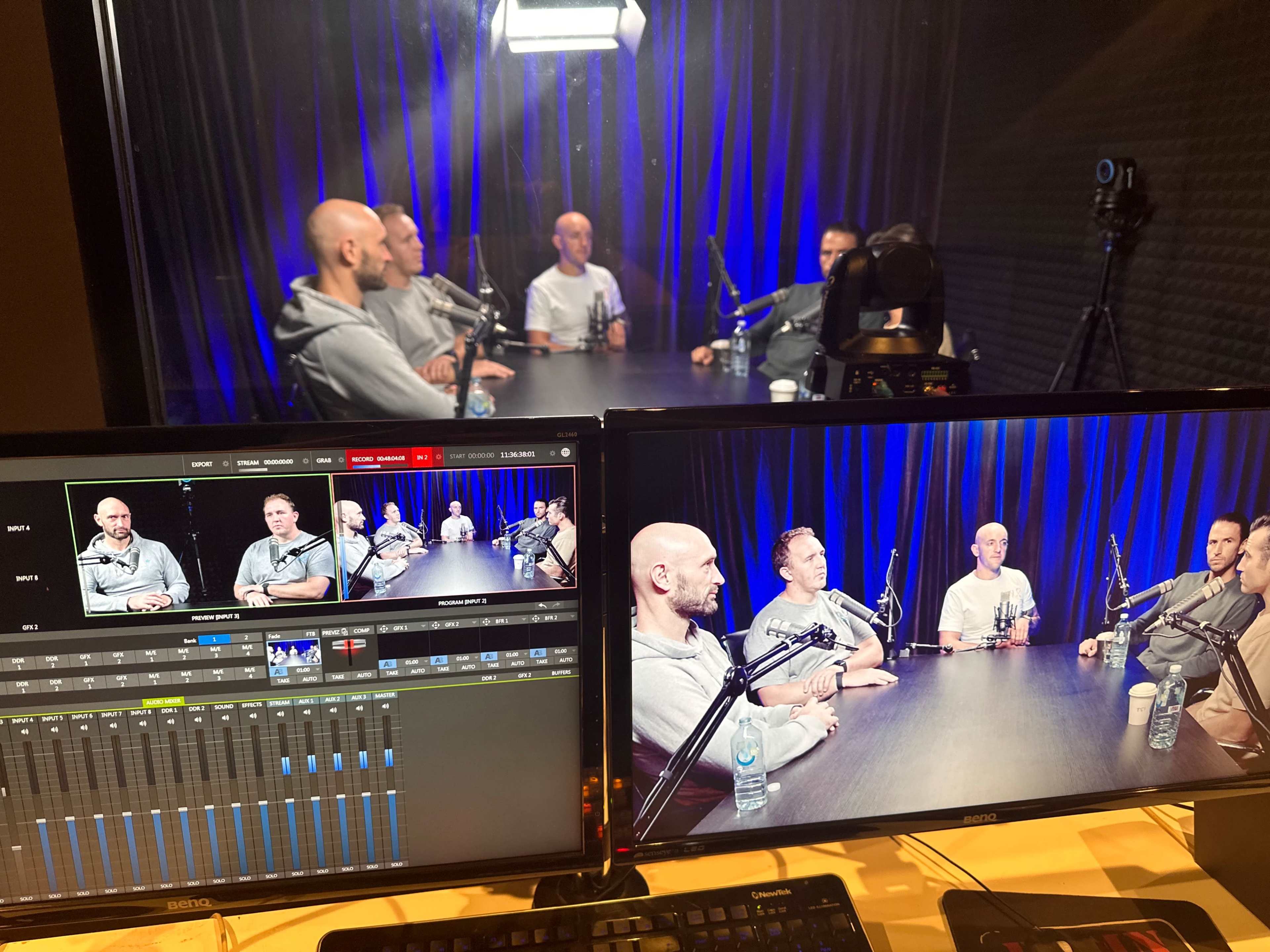 The image shows a video recording setup with multiple monitors displaying a group of five individuals seated at a table, illuminated by overhead lighting in a studio environment.