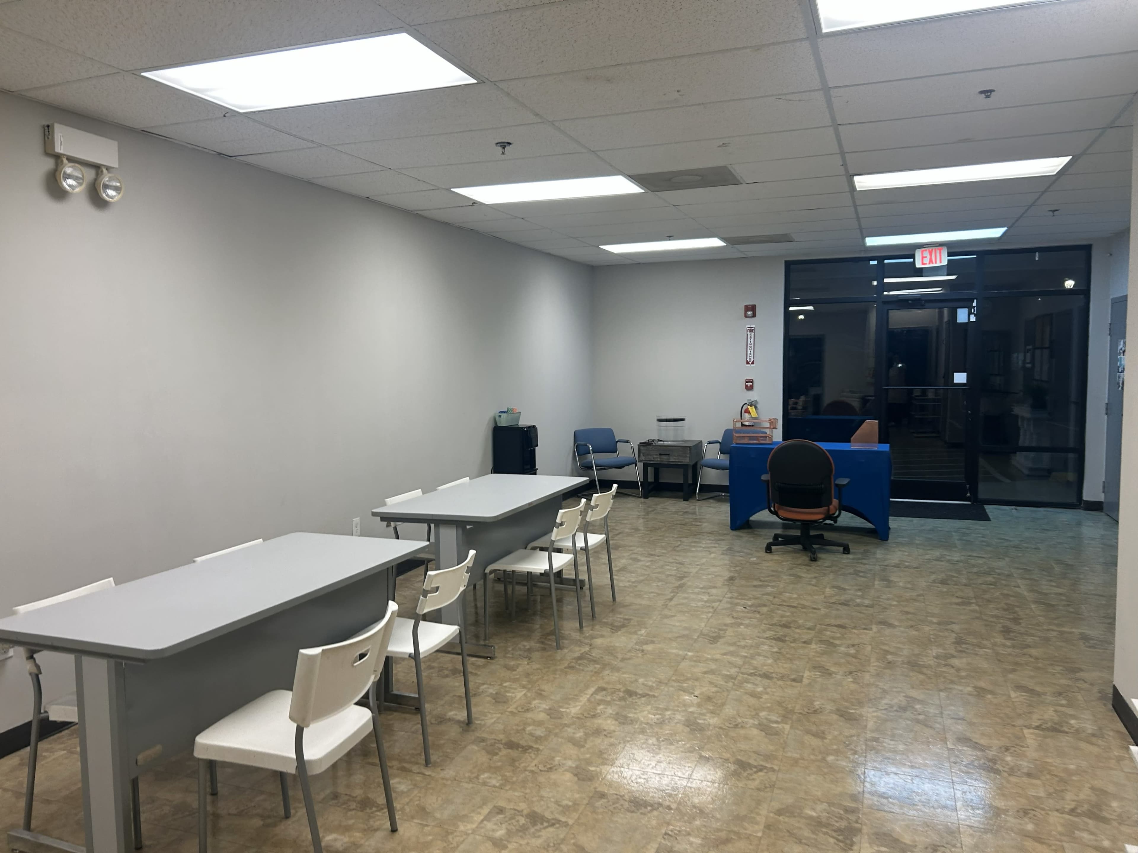 The image shows a spacious, empty room with tables and chairs on one side and a reception or office area on the other, leading to a glass entrance door.