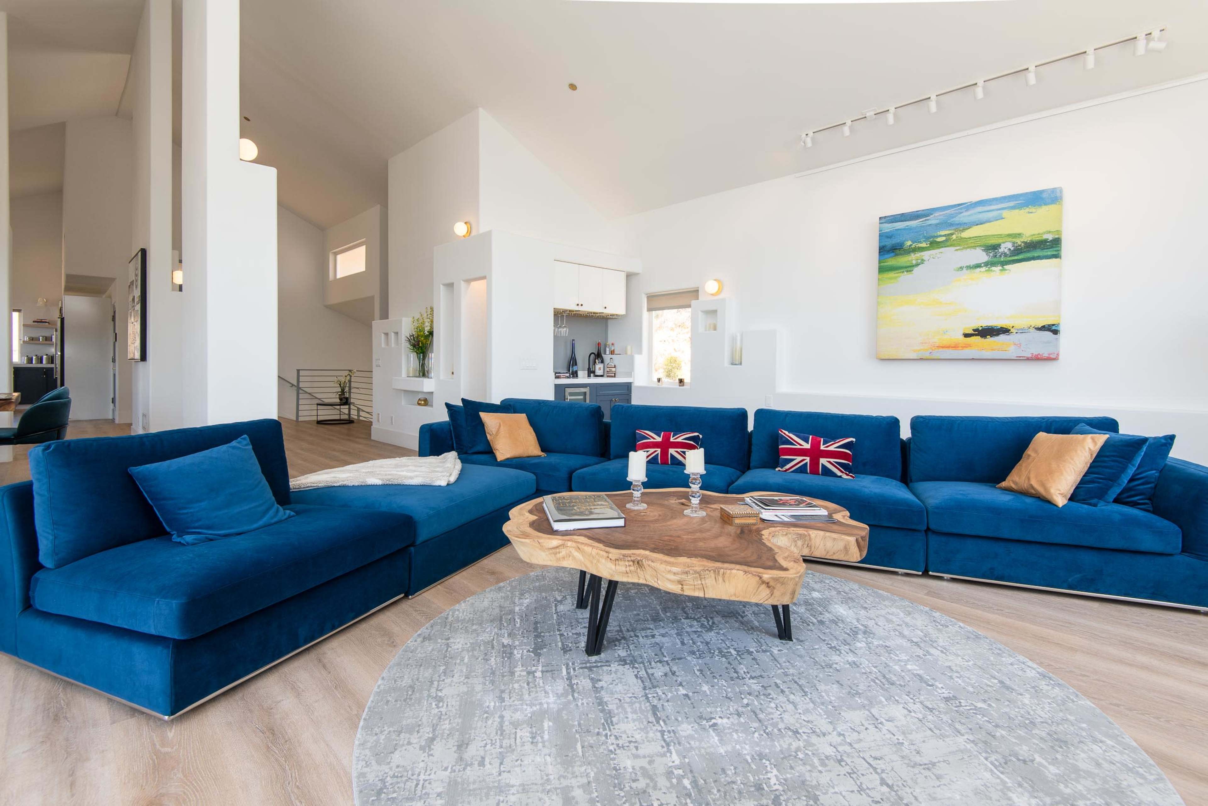 A spacious living room with a large blue sectional sofa, a wooden coffee table with a unique shape, and a vibrant landscape painting on the wall.