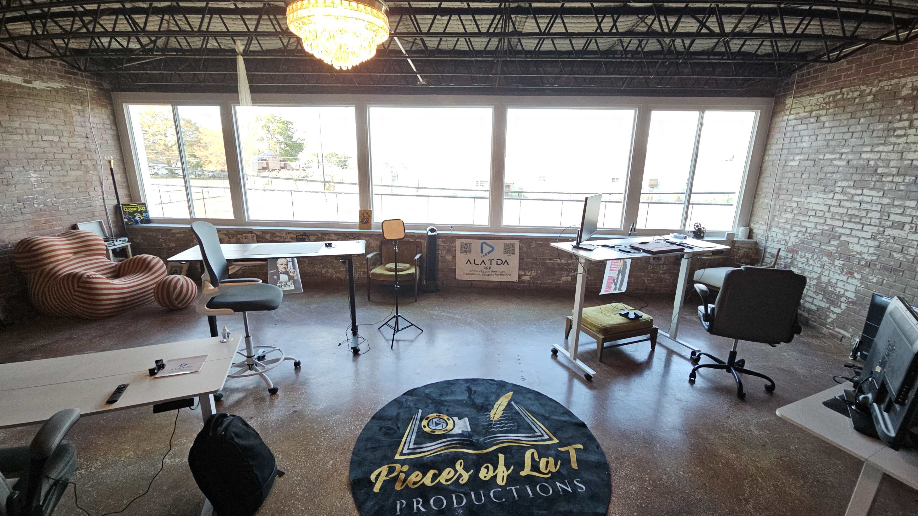 The image shows a spacious office with large windows, exposed brick walls, various workstations, and a round rug displaying the words "Pieces of La T Productions."