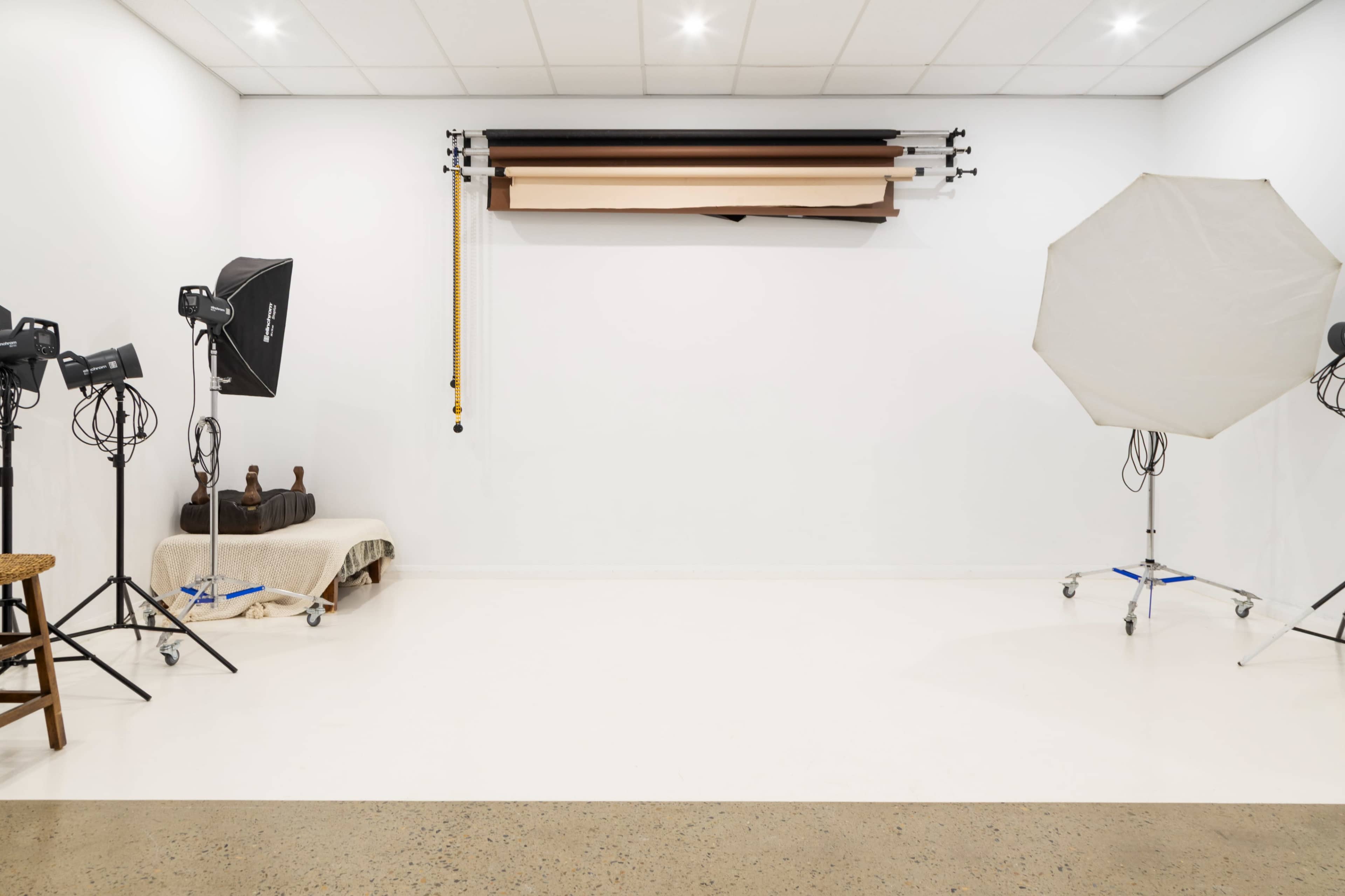 The image shows a photography studio with two lighting setups, a backdrop storage system, and a small seating area.