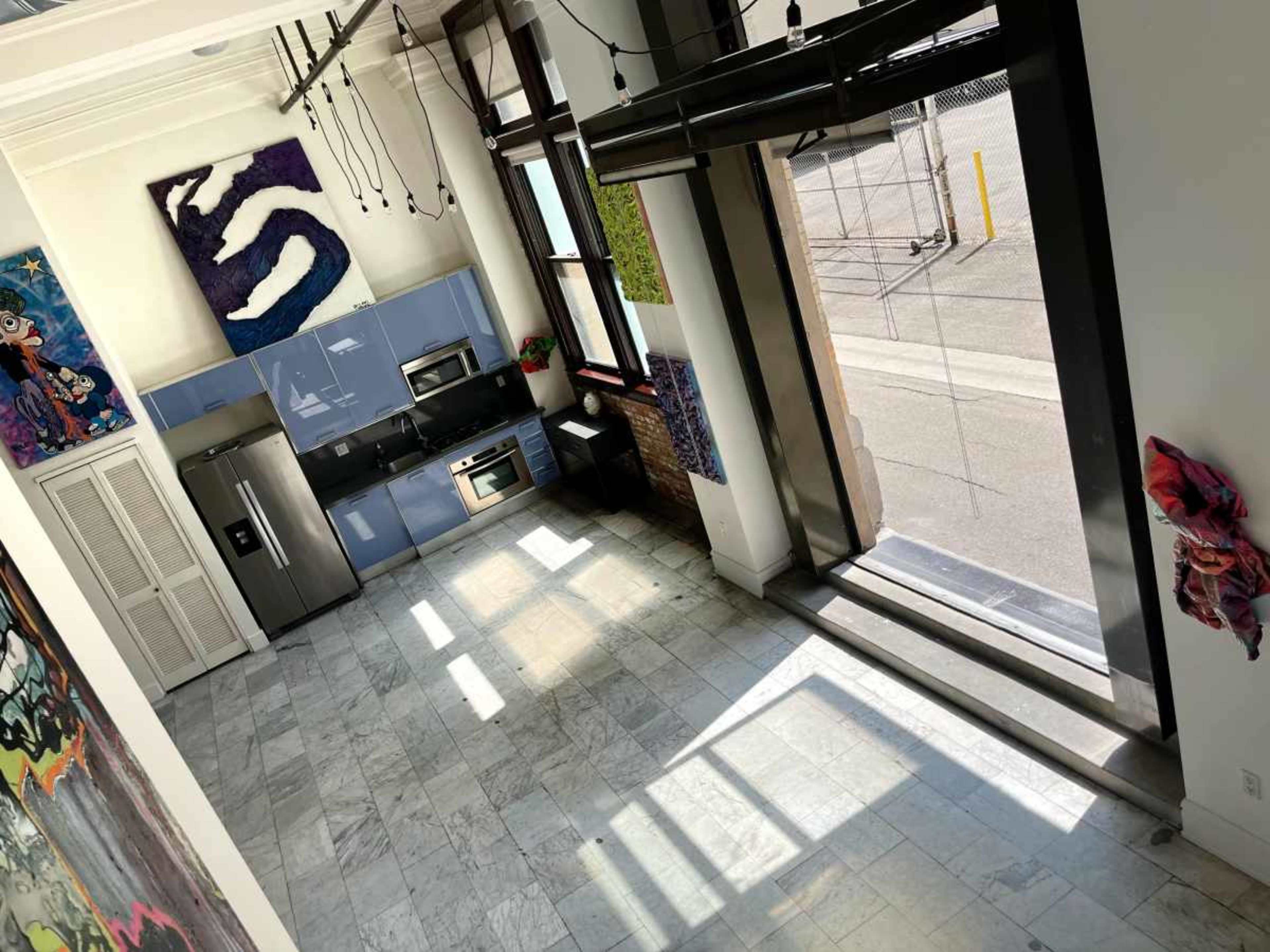 The image shows a modern kitchen area with blue cabinets and stainless steel appliances, featuring large windows that allow natural light to enter a spacious, minimally decorated room with marble flooring.