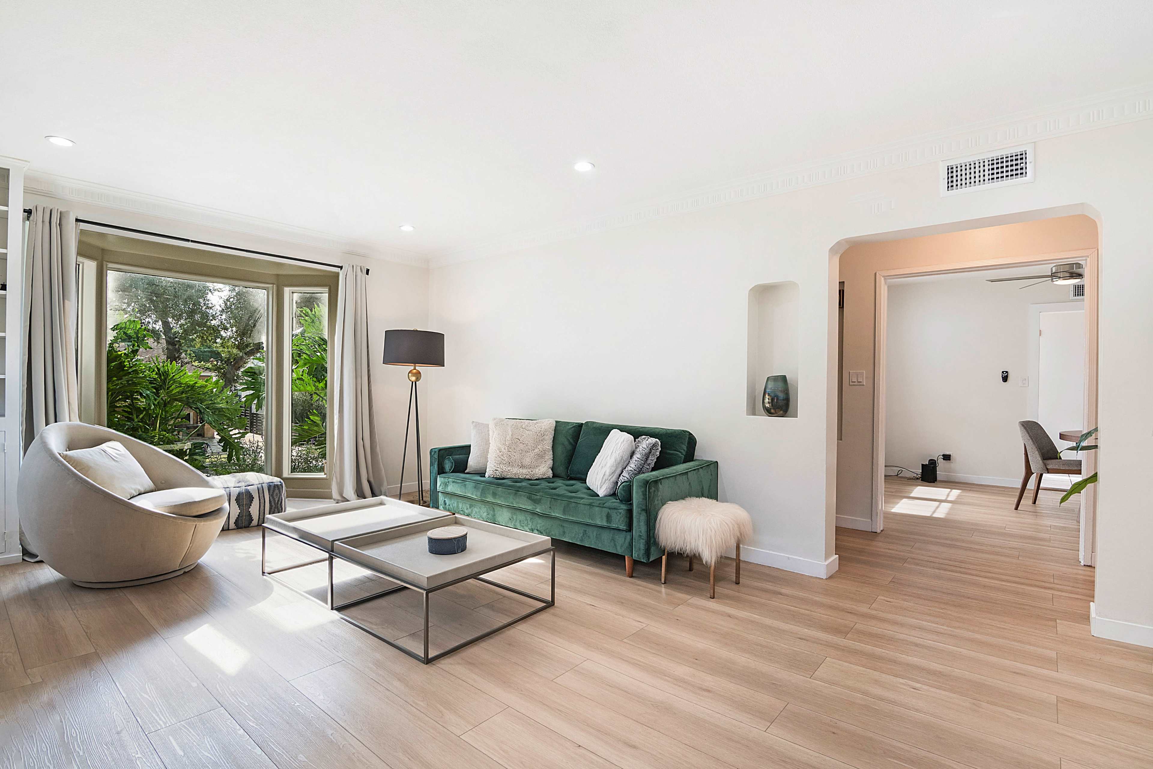 The room features a green sofa, a round chair, and a coffee table, with large windows showcasing greenery outside.