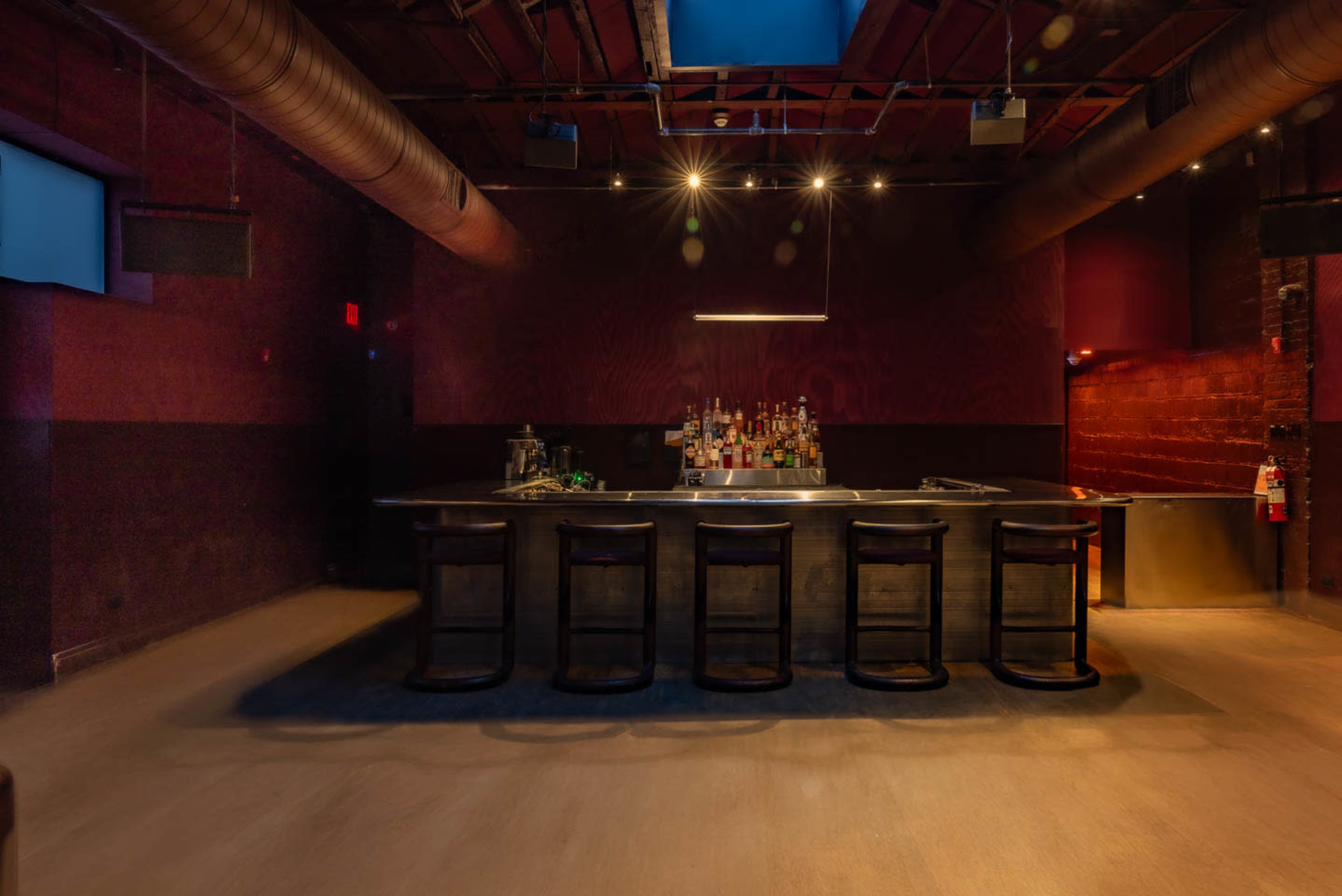 The image shows a dimly lit bar with a long countertop, several bar stools in front, and shelves of liquor bottles in the background.