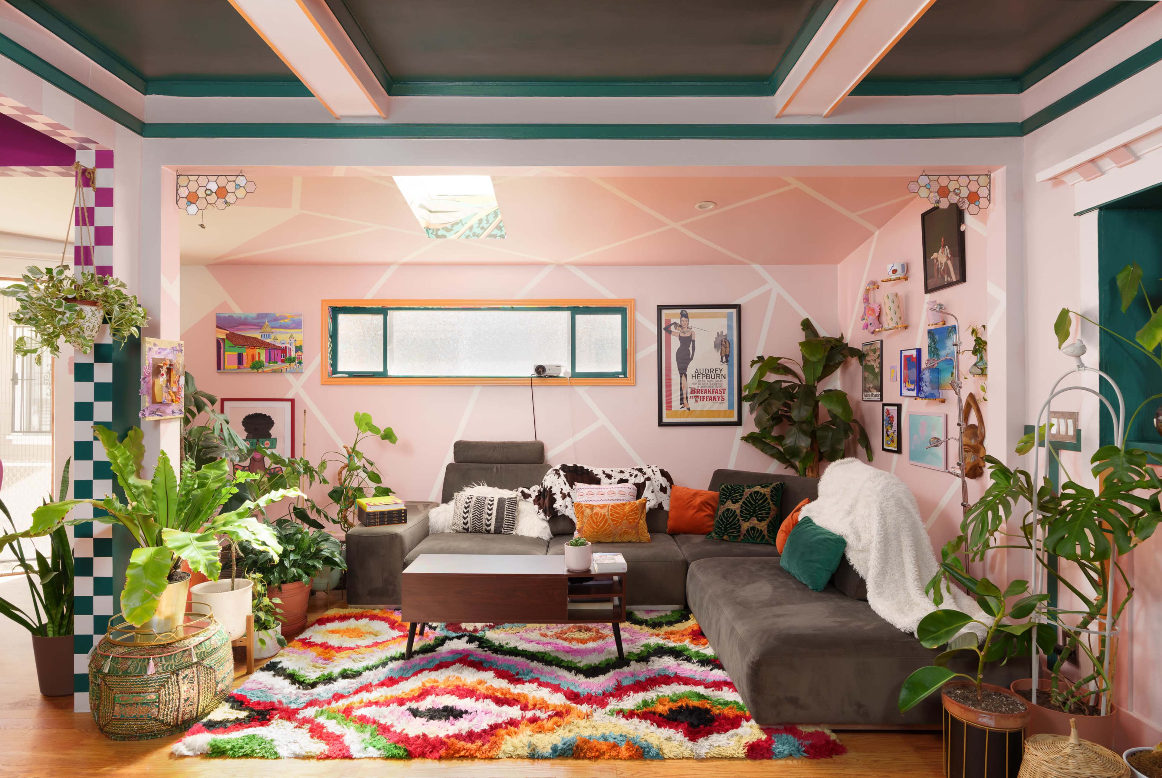 The image shows a colorful living room with a gray sectional sofa, a vibrant rug, and various plants arranged around the space.