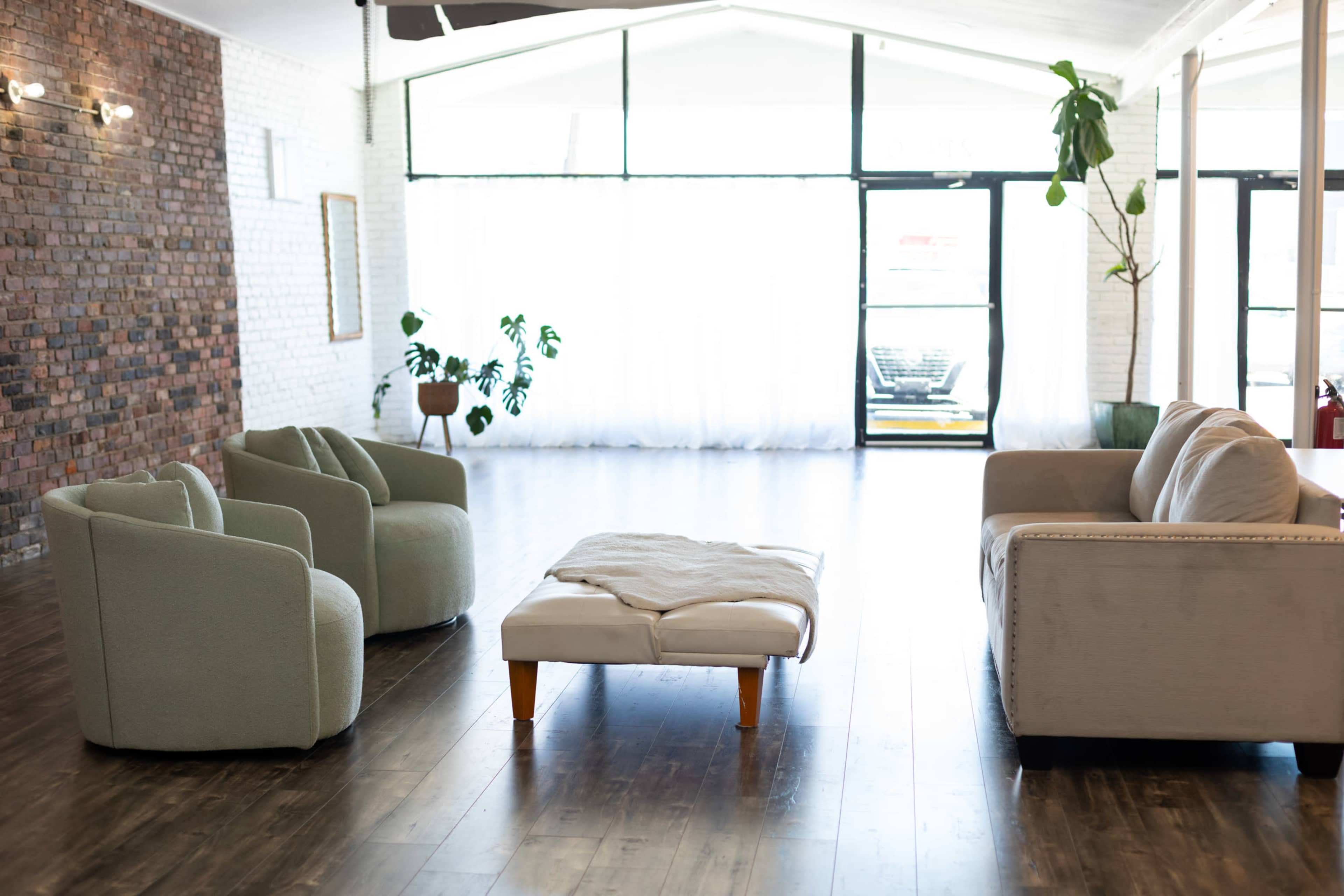 The image shows a spacious room with a brick wall, three green chairs, a light-colored sofa, and a wooden coffee table in the center.