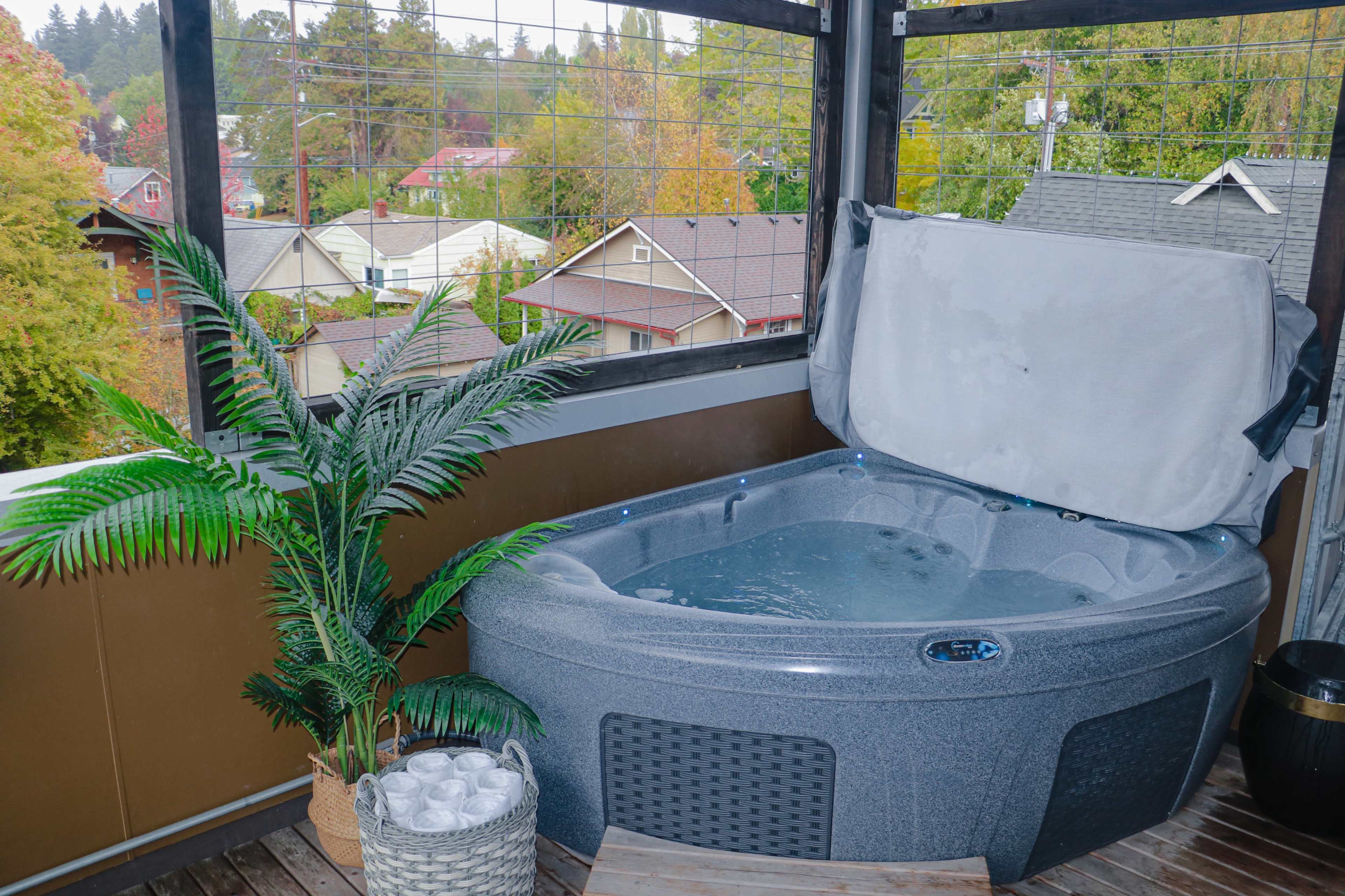 A hot tub is positioned on a wooden deck, surrounded by a potted plant and overlooking a neighborhood with houses and trees.