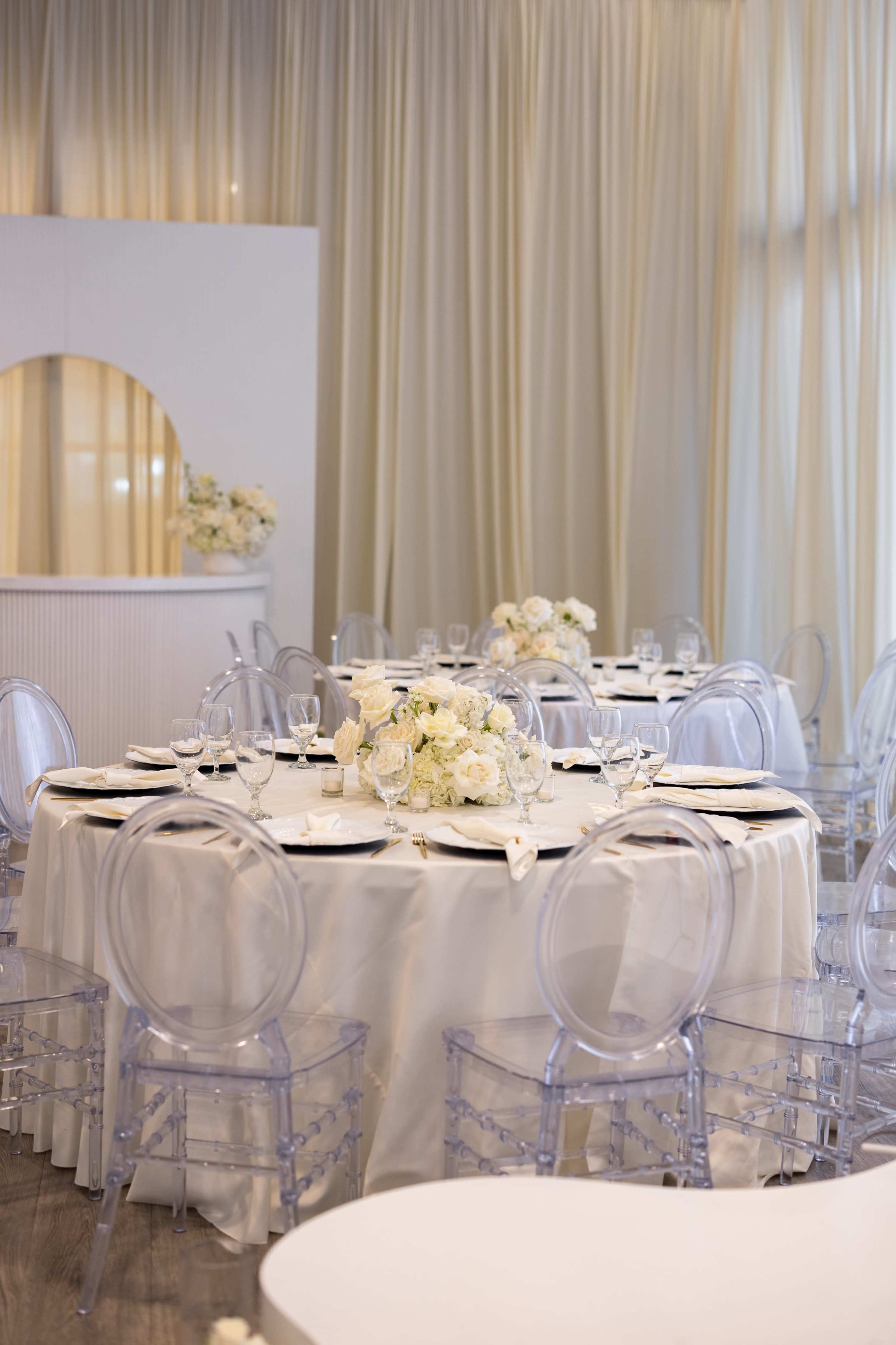 A elegantly set dining area features round tables with white tablecloths, clear acrylic chairs, and floral centerpieces of white roses.