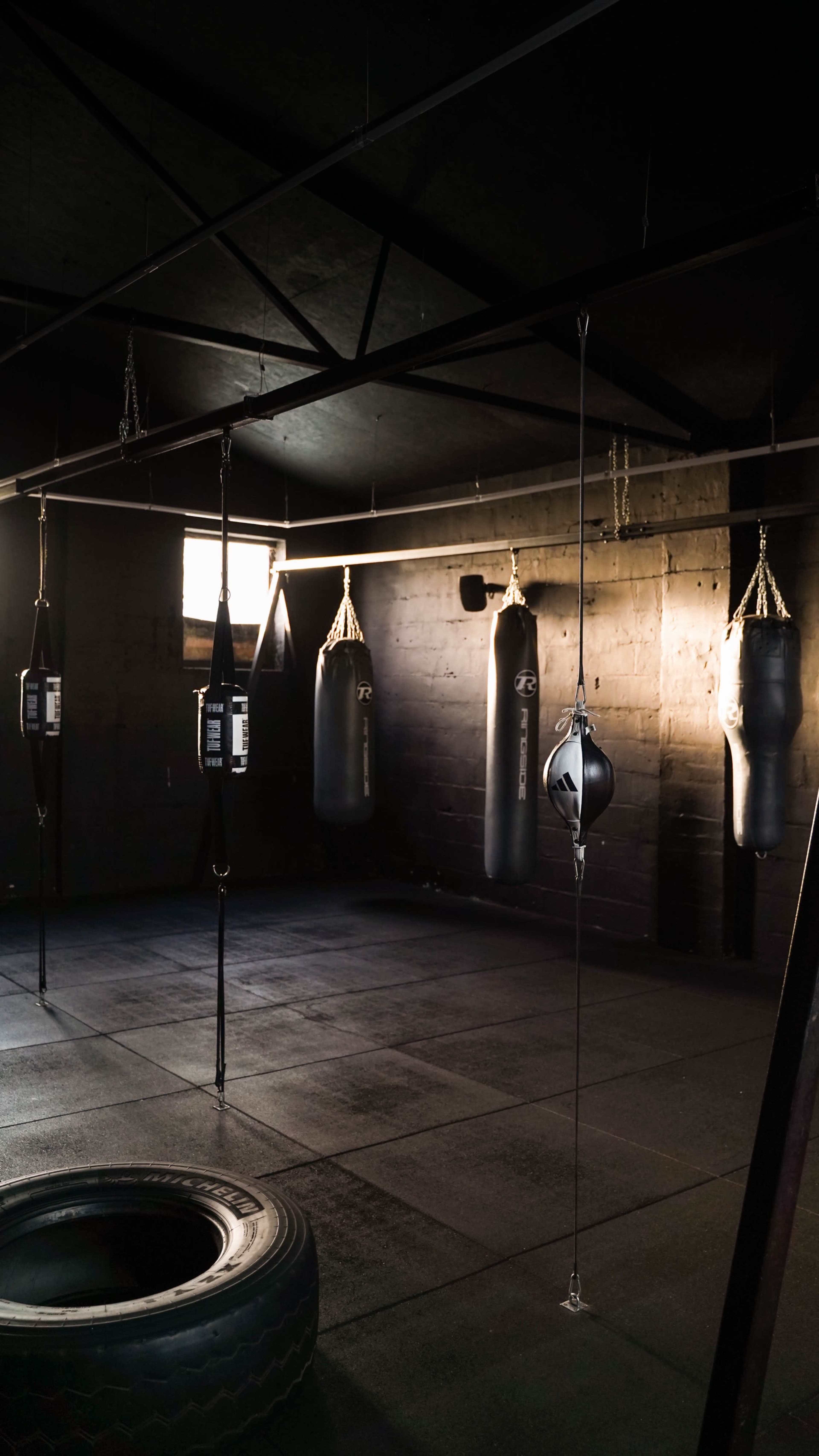 The image shows a dimly lit gym with several hanging boxing bags and a large tire on the floor.