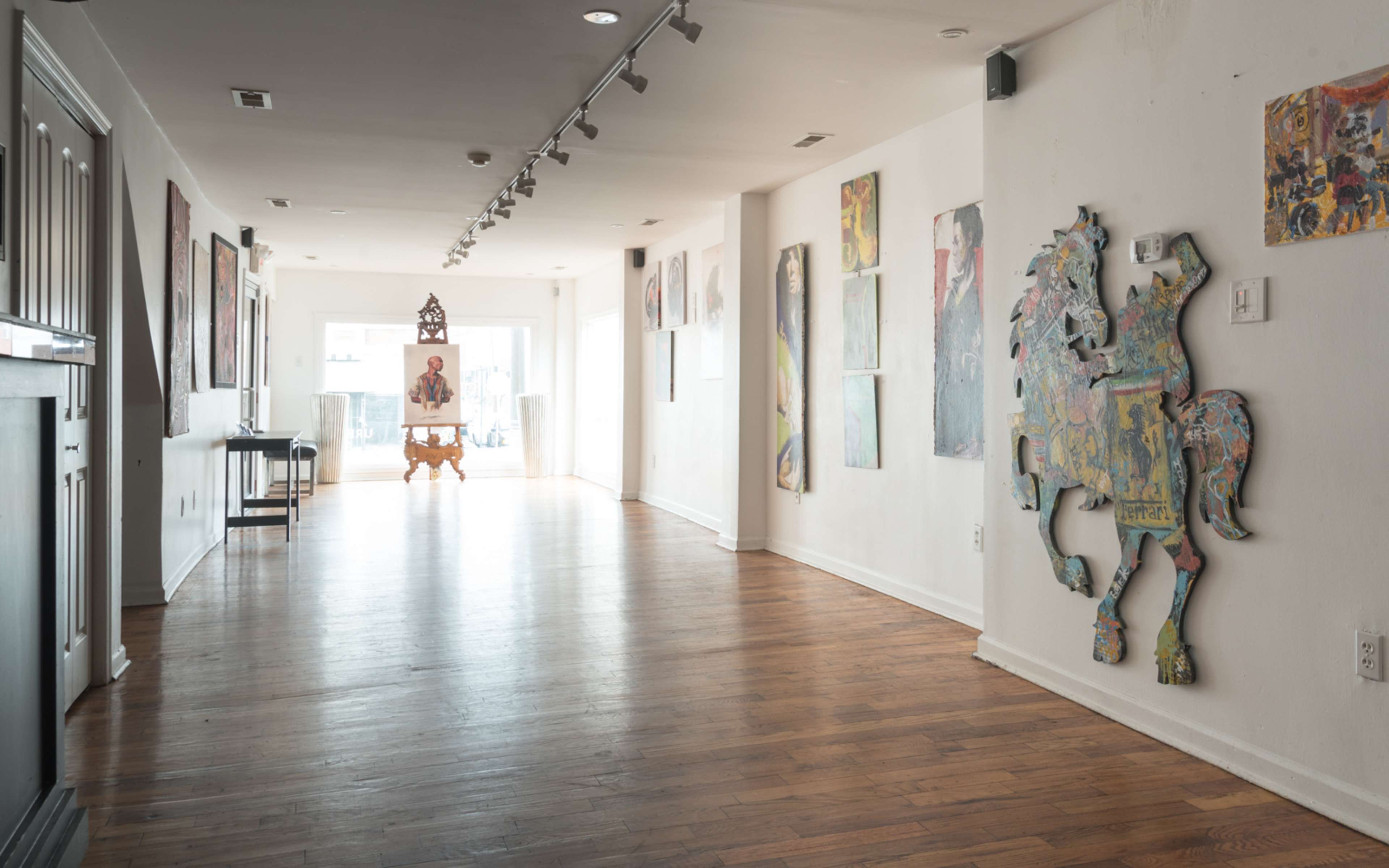 A spacious, well-lit gallery featuring various artworks on the walls and a wooden sculpture at the far end.