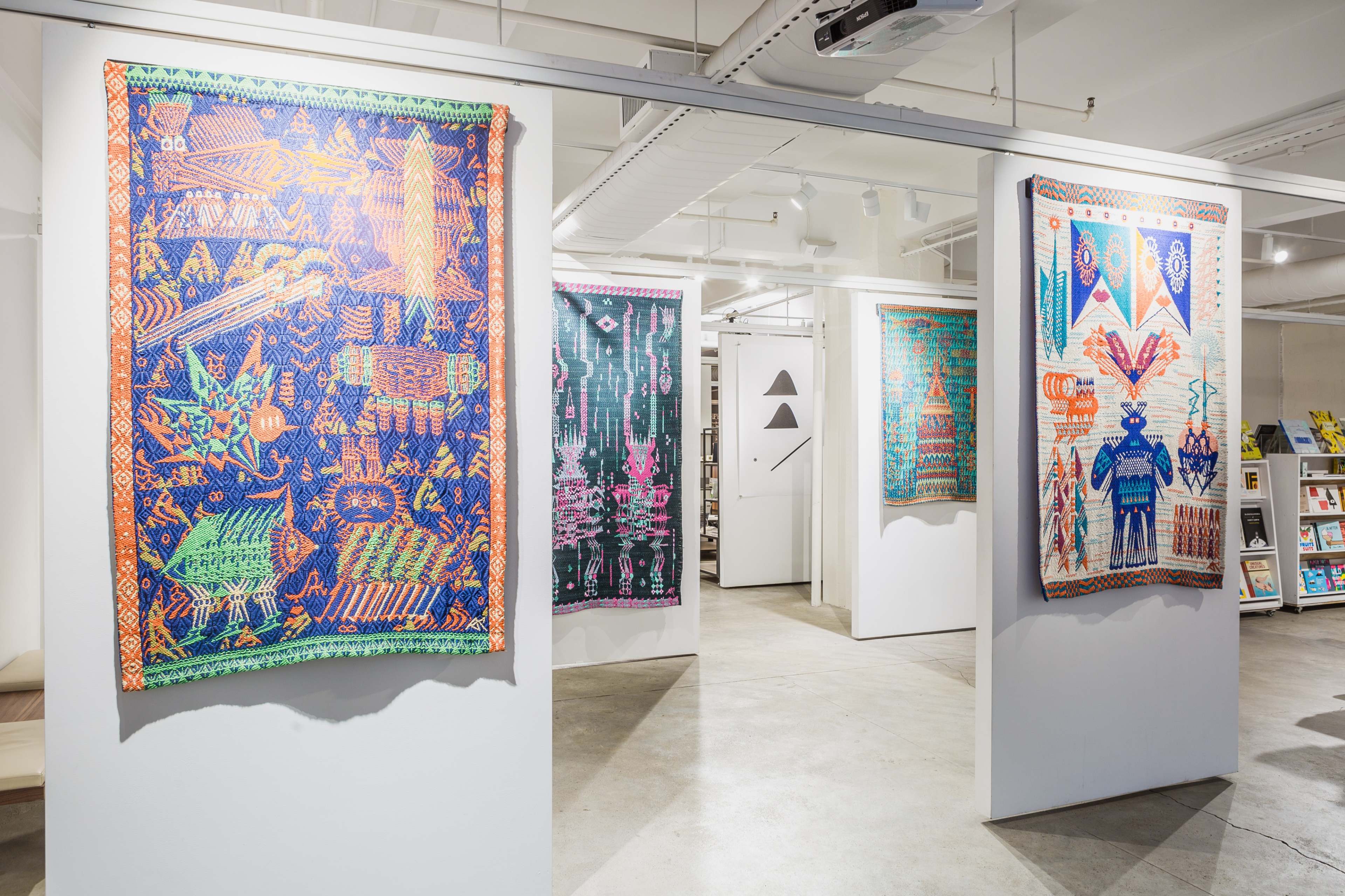 A gallery space featuring colorful, patterned wall hangings displayed on white walls, with bookshelves visible in the background.