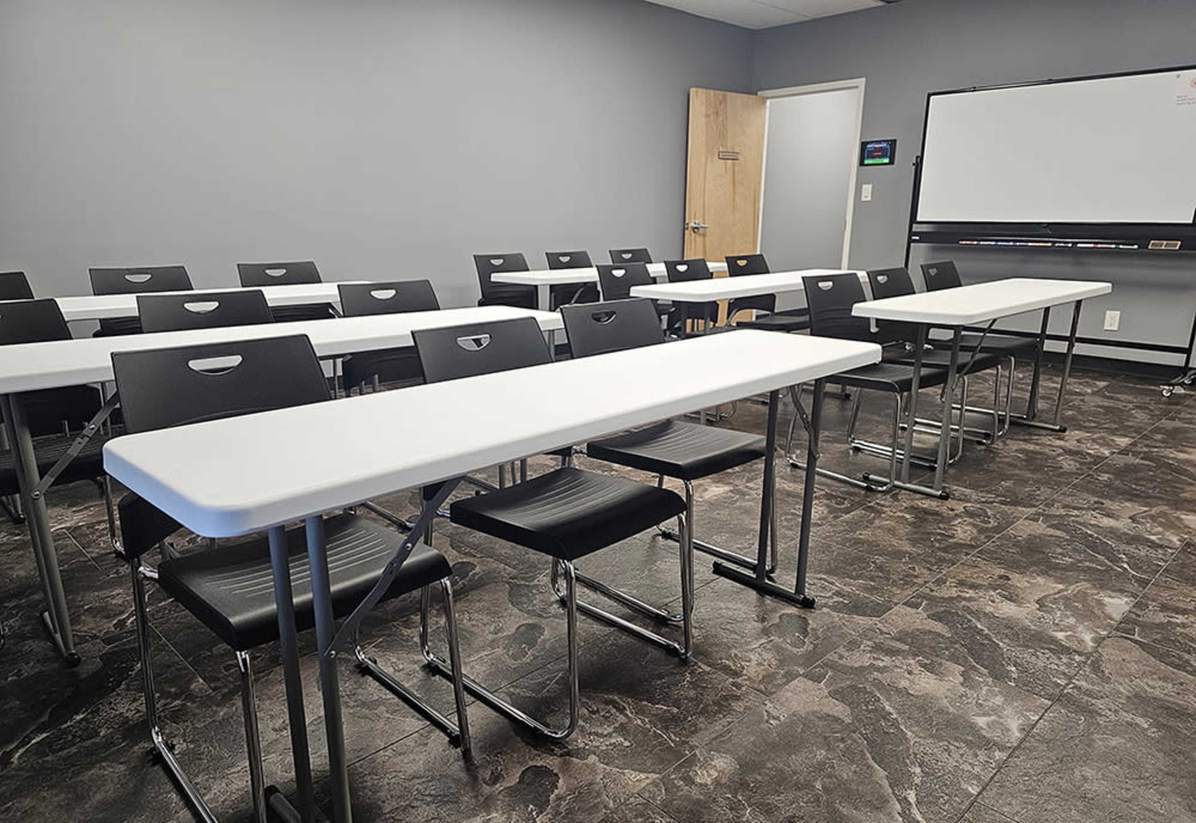 A classroom is set up with several rows of white tables and black chairs arranged neatly against a gray wall, with a screen visible at the front.