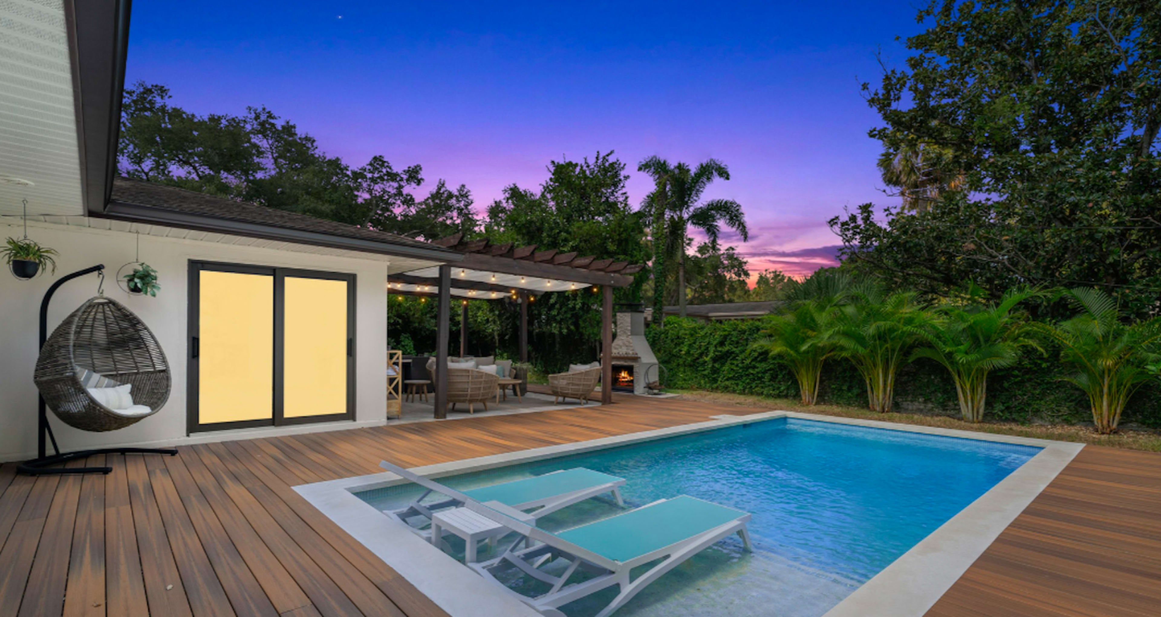 A backyard with a swimming pool, wooden deck, and outdoor seating area under a pergola, set against a sunset sky.