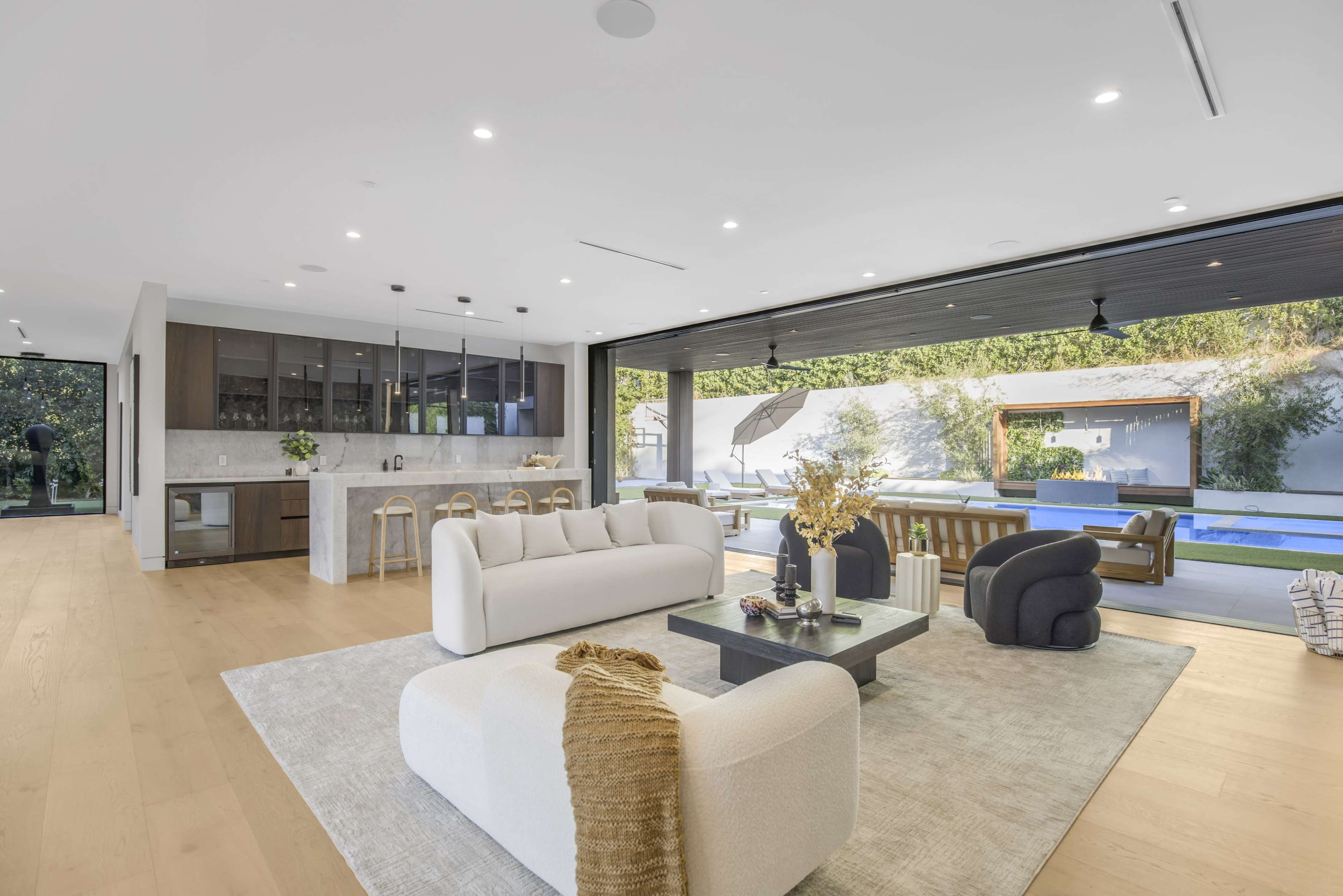 The image shows a modern living space featuring an open layout with a large sectional sofa, a coffee table, and a kitchen area with bar stools, leading to an outdoor pool area.