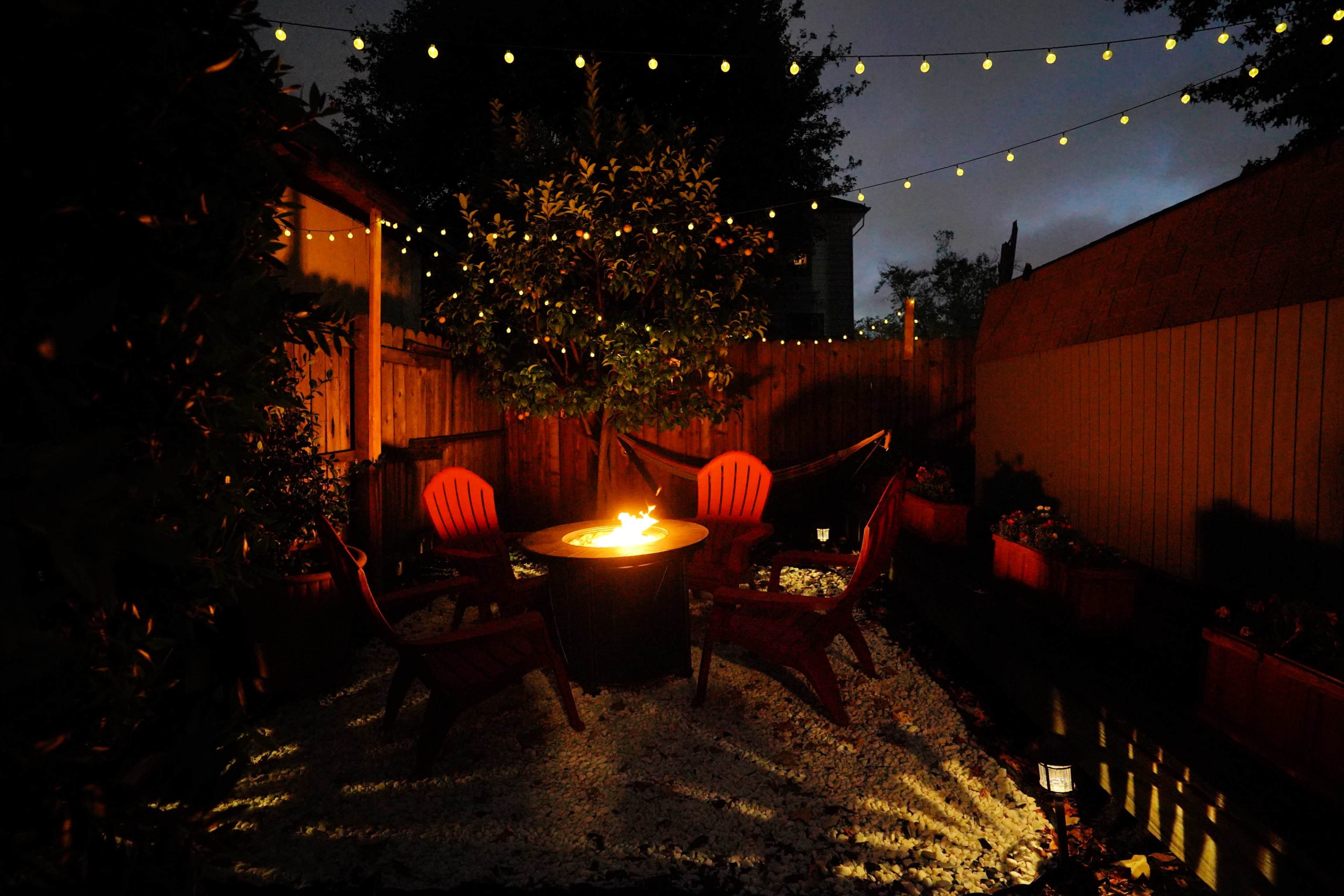 A circular fire pit surrounded by four red chairs is illuminated under string lights in a backyard garden at night.