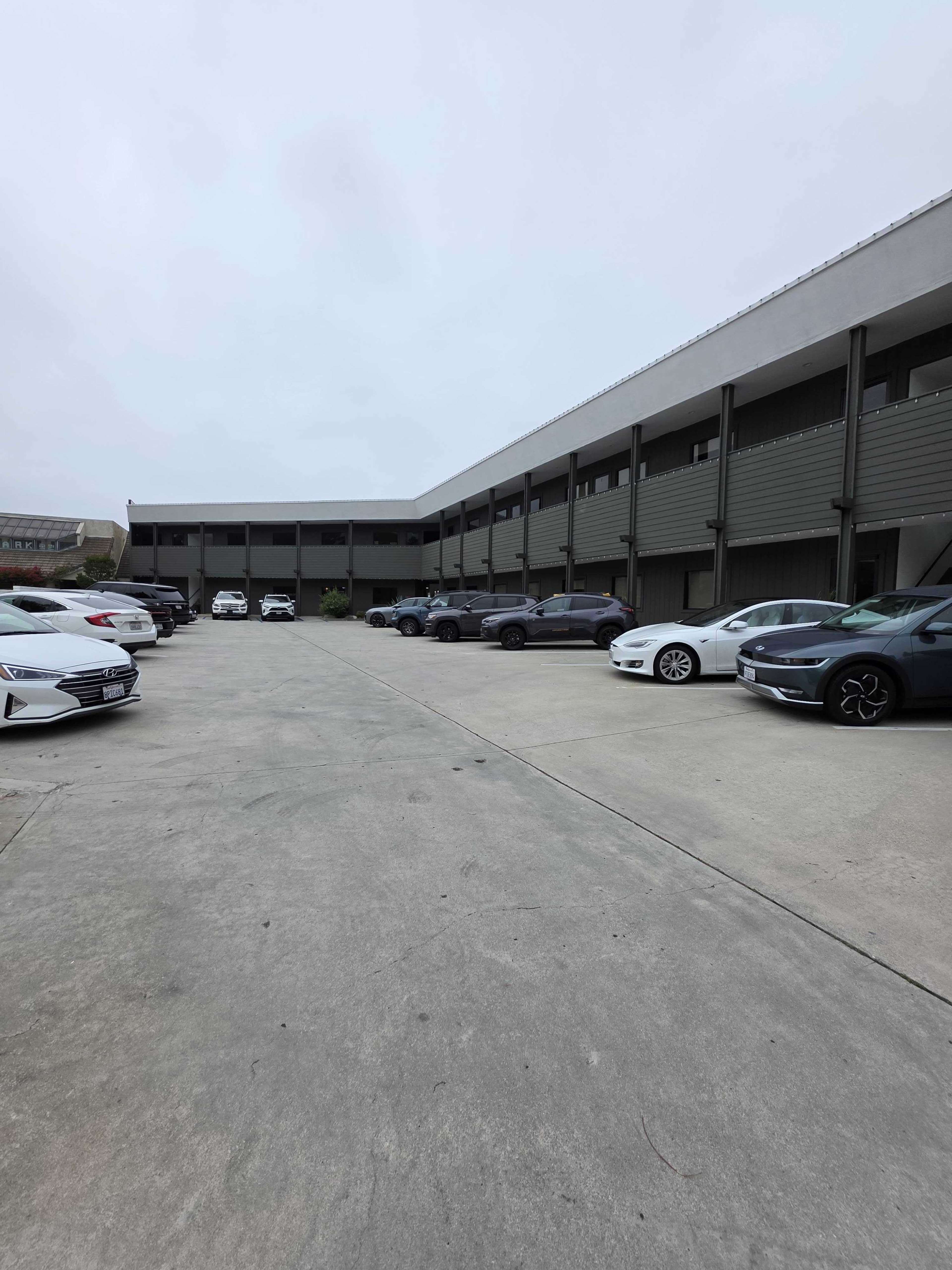 The image shows a parking lot with several cars arranged along both sides of a concrete lane beside a two-story building.