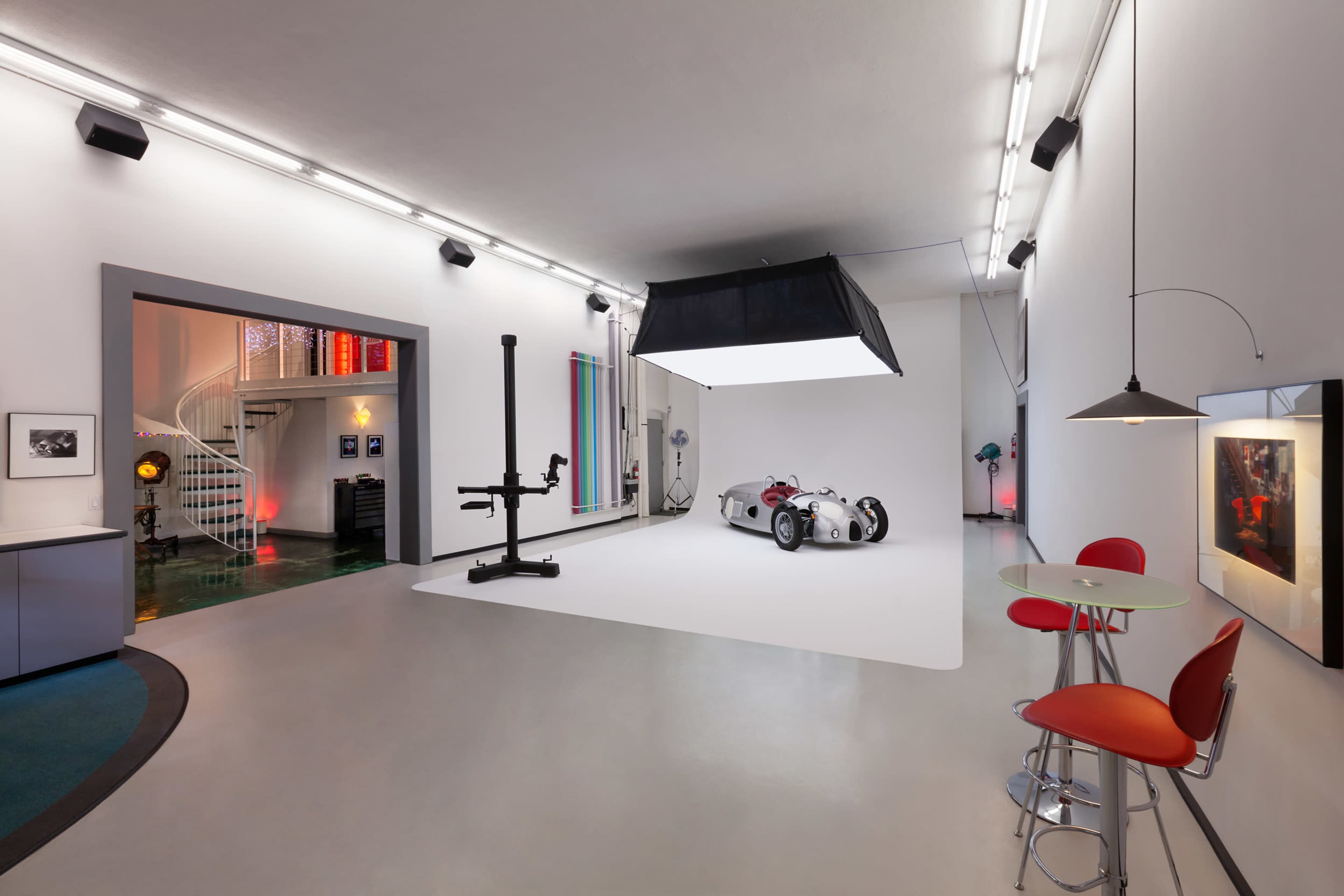 A modern studio space featuring a vintage sports car on a white backdrop, illuminated by large overhead lighting, with a minimalist seating area and artistic decor on the walls.