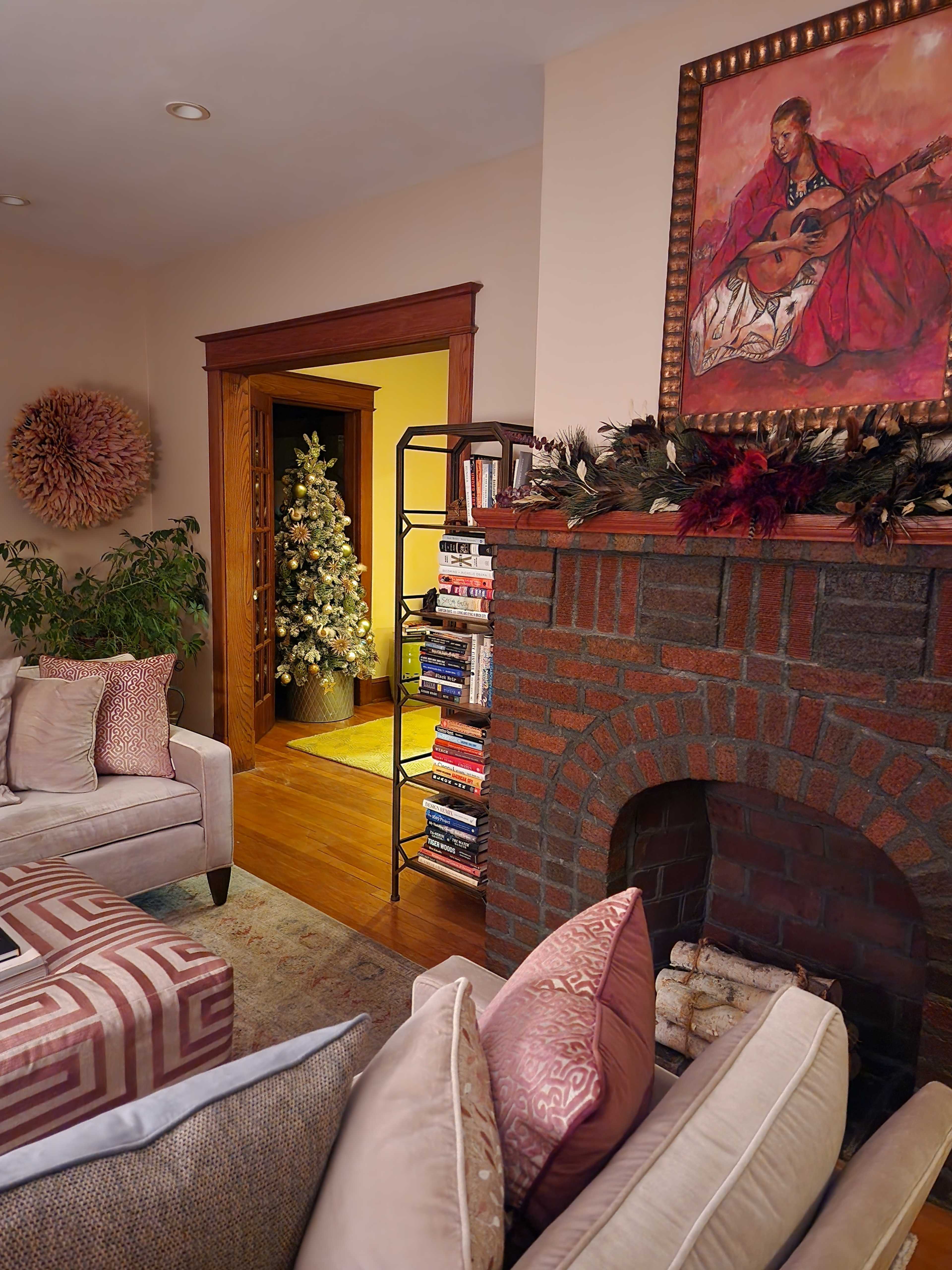The image shows a cozy living room featuring a couch with decorative pillows, a brick fireplace, a bookcase filled with books, and a beautifully decorated Christmas tree in the adjacent room.