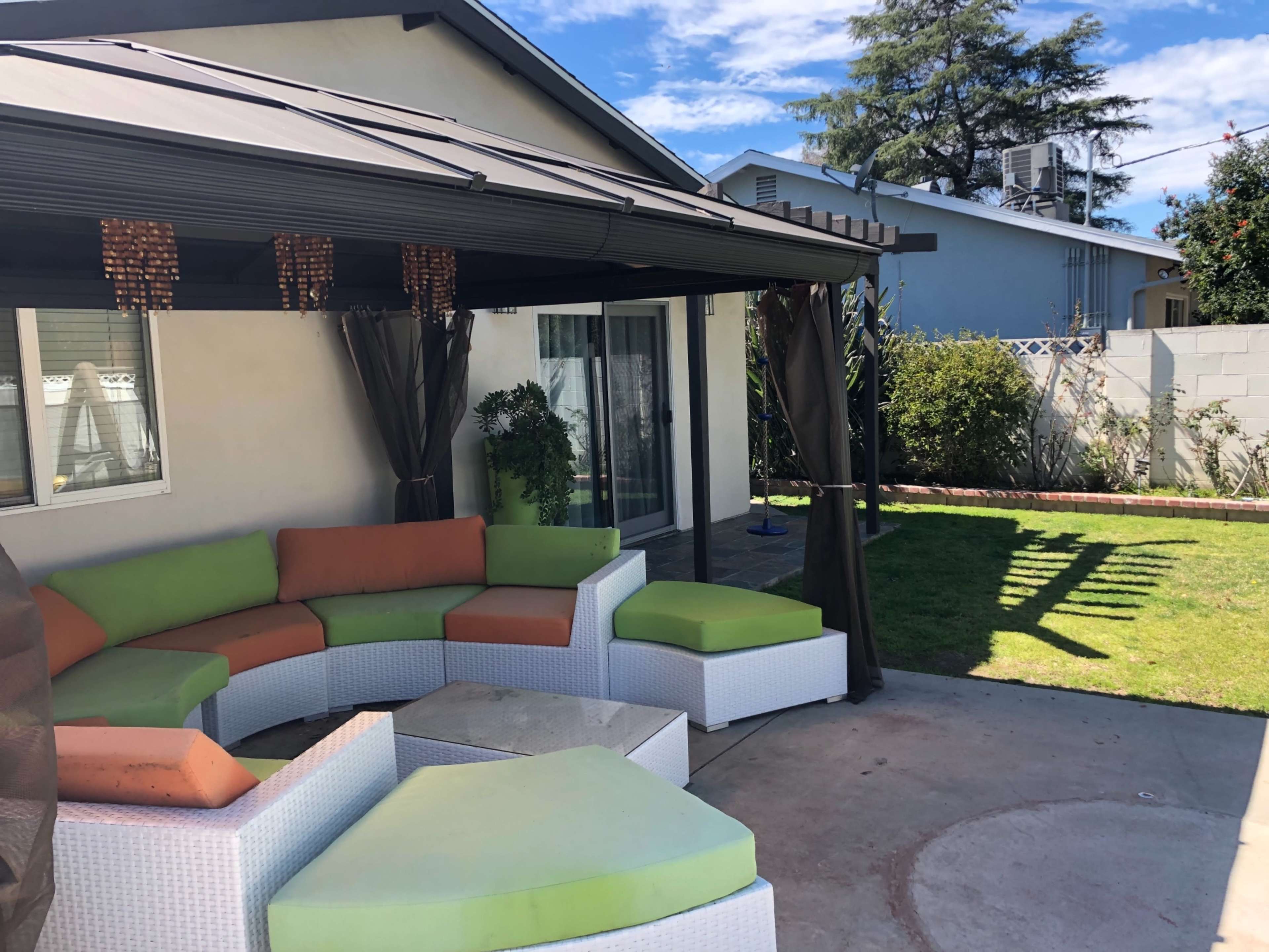 A covered outdoor seating area with a curved sectional couch in green and orange sits in a backyard with a grassy lawn and a wall.